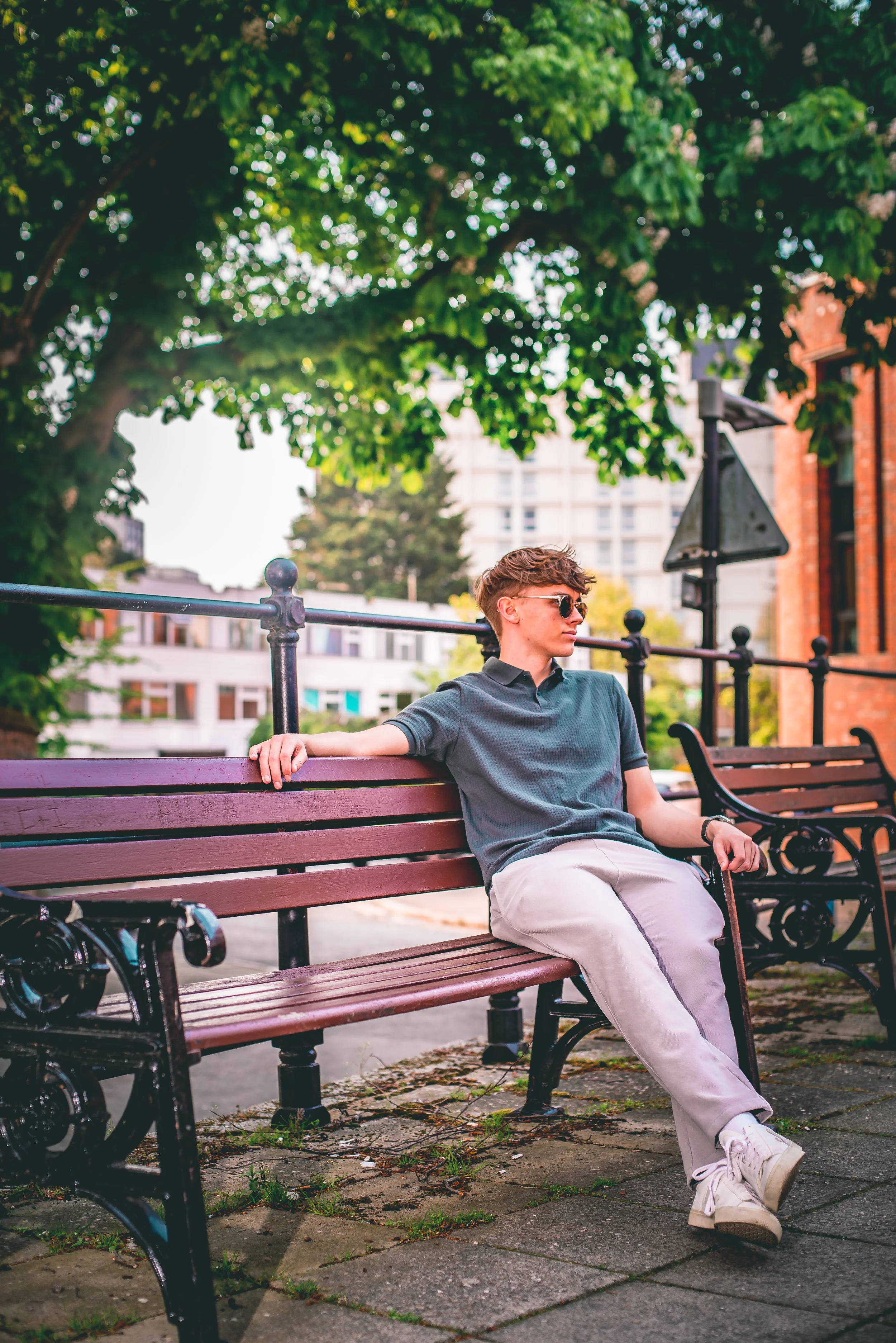 A young man with short brown hair, wearing sunglasses, a gray polo shirt, and beige pants, is sitting on a wooden park bench with black metal armrests and legs. He is relaxed, with his right arm resting on the backrest, and his legs crossed at the an