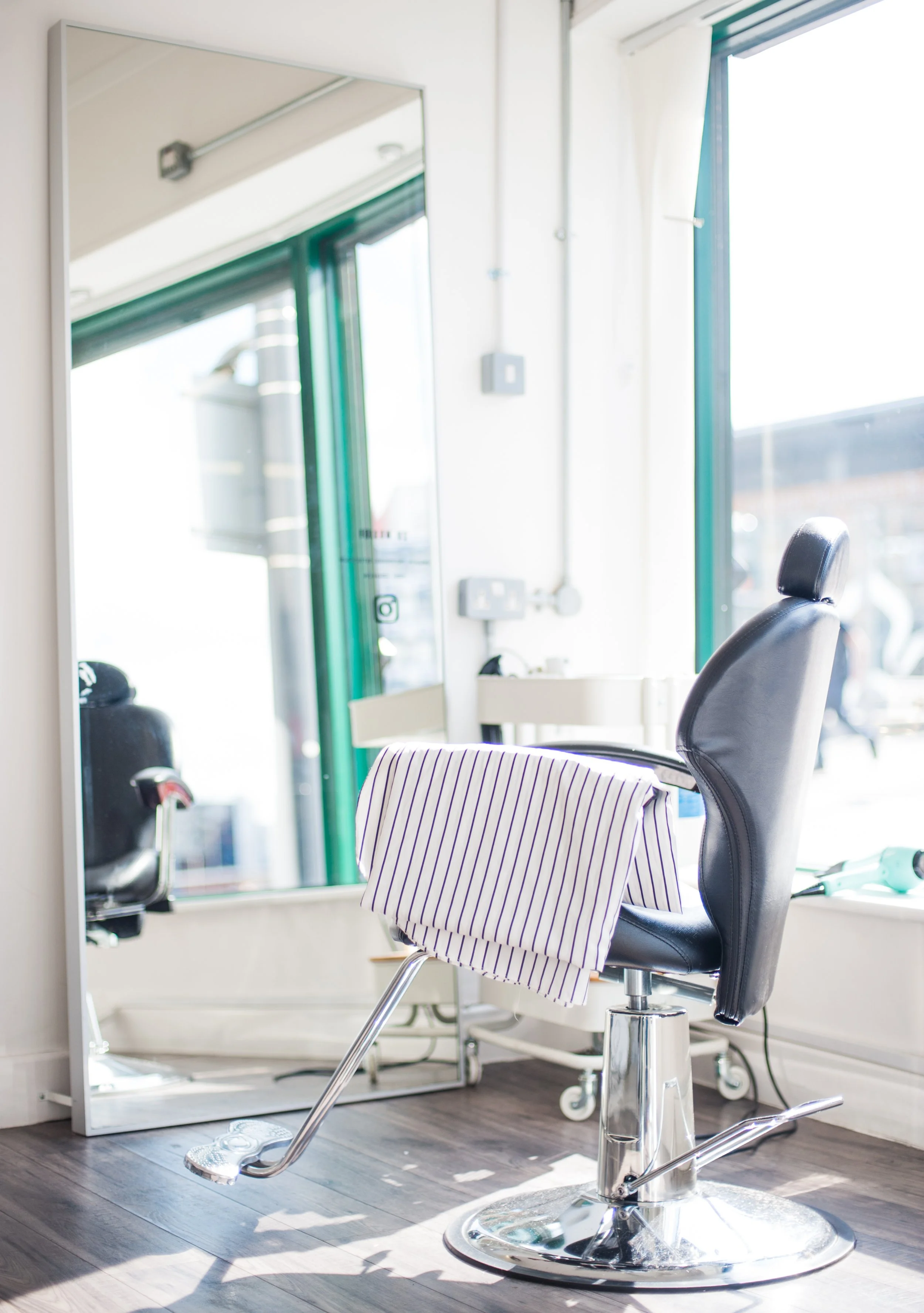 Haircutting chair in a salon with a striped cloth draped over it, in front of a large mirror, near a window with sunlight streaming in.