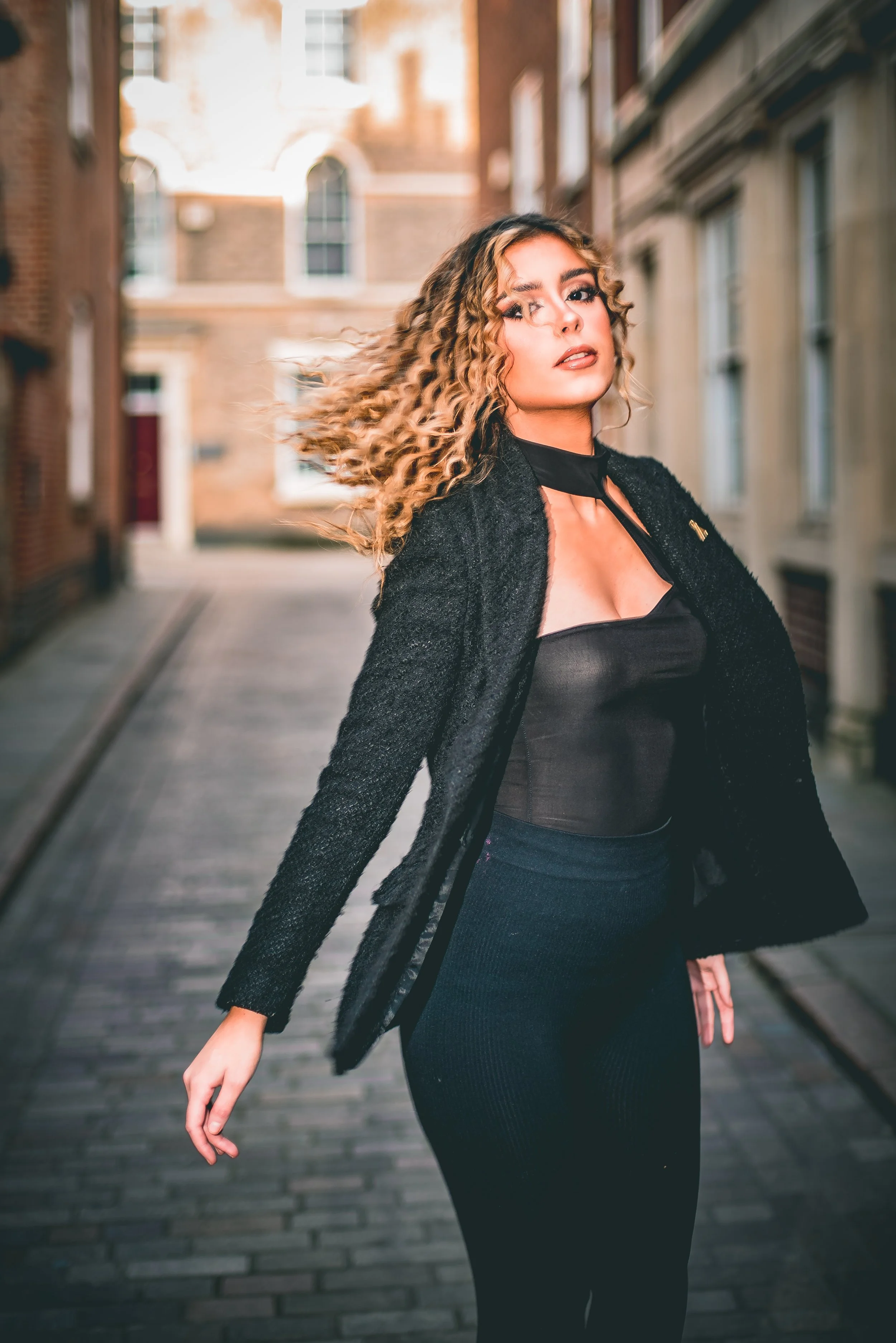 A woman with curly blonde hair walking on a city street, wearing a black top, black skirt, and a black coat, with buildings in the background.