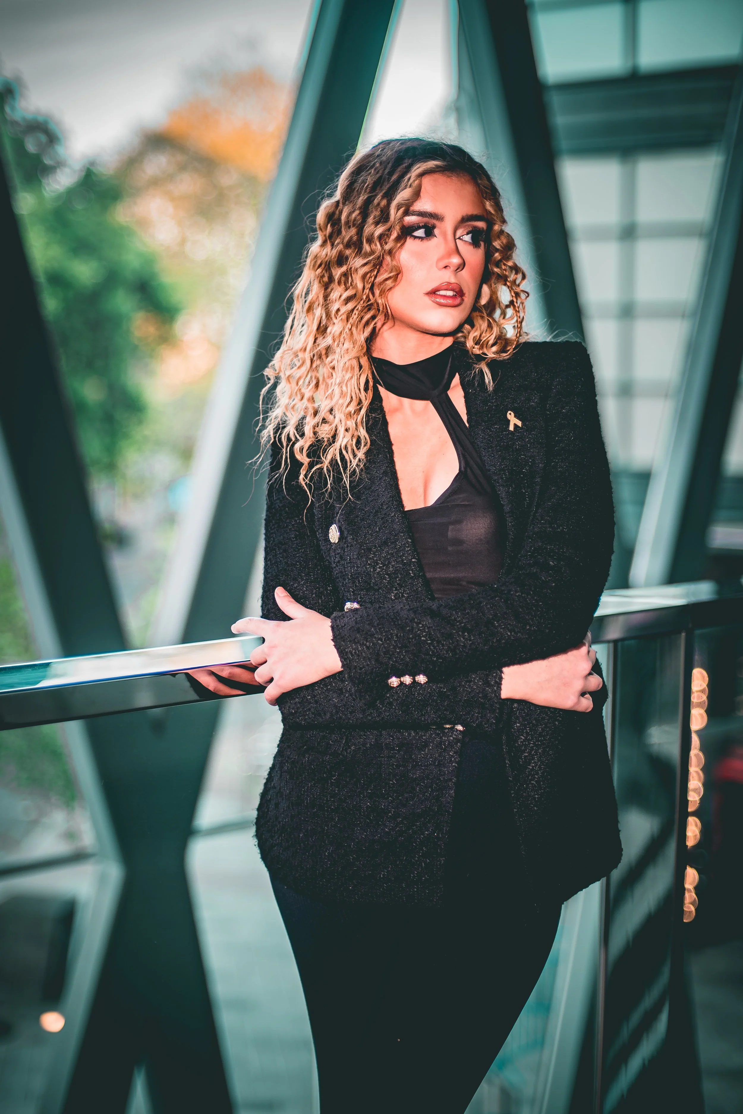 A woman with curly blonde hair looking to the side, wearing a black blazer and a pin, standing in a glass-walled building.