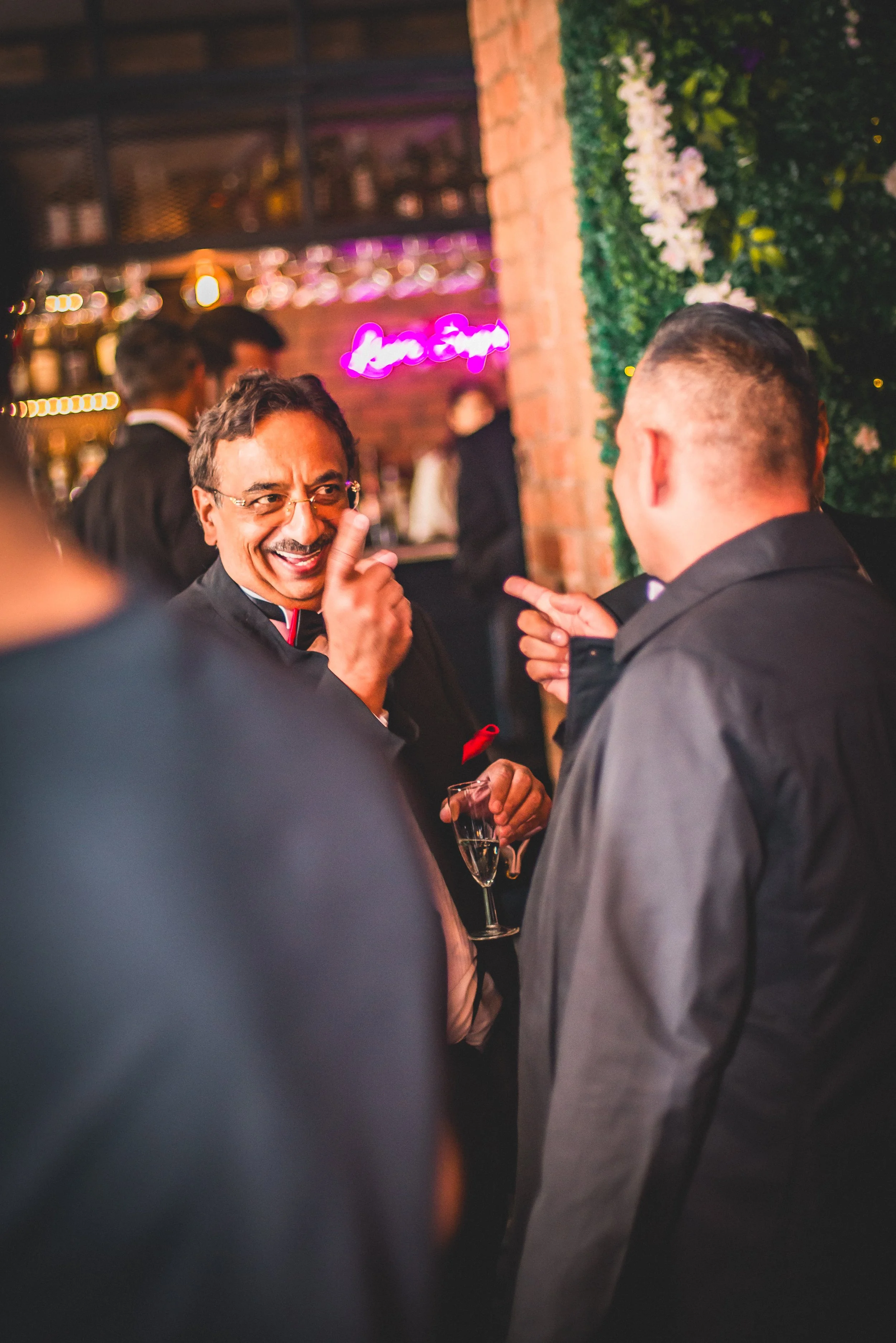 Two men in suits and ties are engaged in conversation in a lively bar or lounge, with one smiling and pointing while holding a glass of champagne.