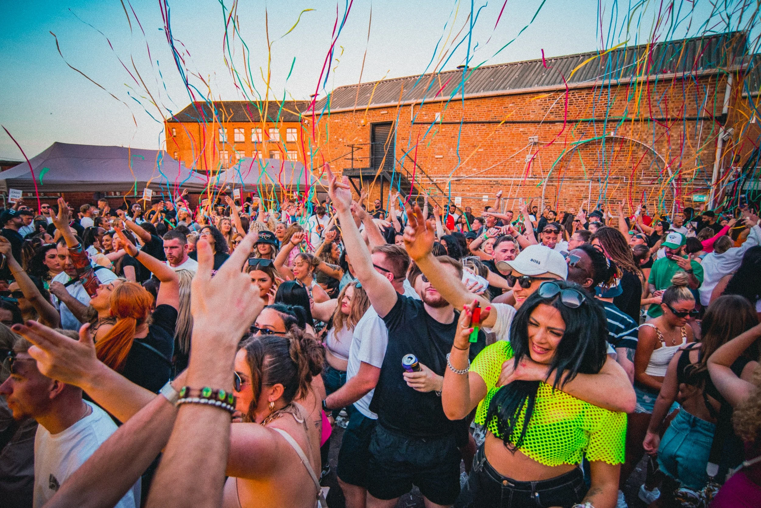 Crowd of people dancing and celebrating outdoors with colorful streamers in the air, brick building in the background.