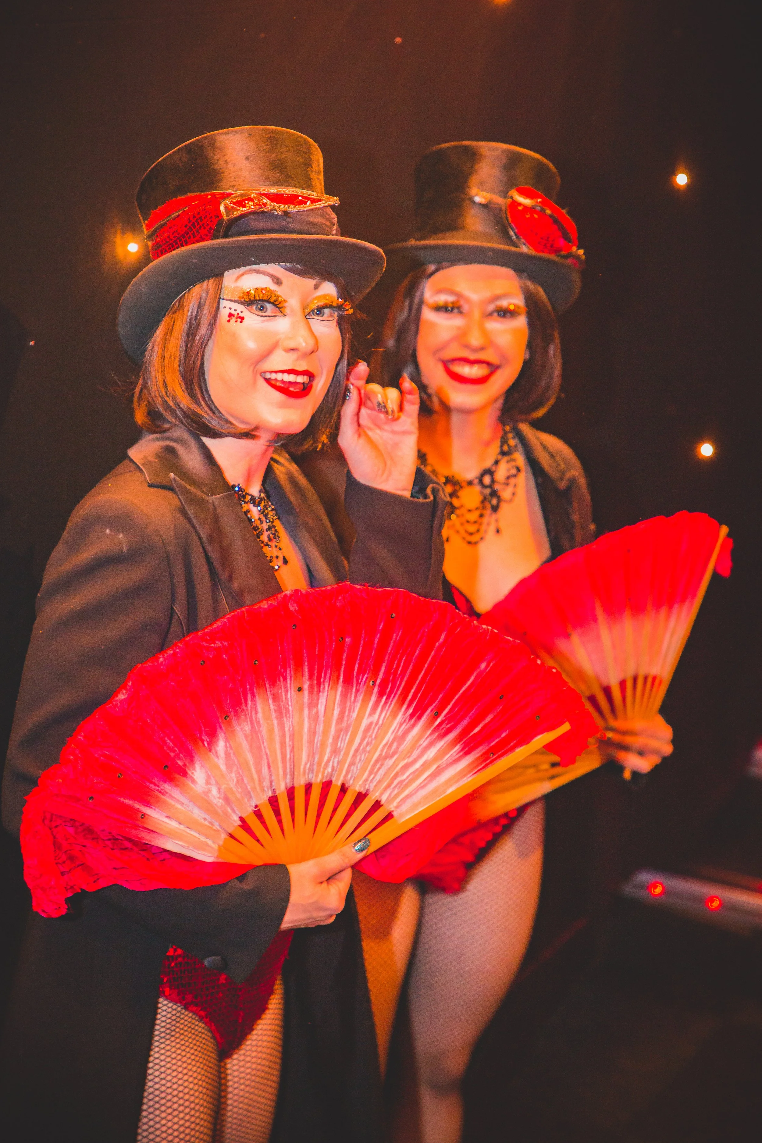 Two women dressed in burlesque costumes holding red fans, wearing black top hats with red accents, with bright makeup, in a dark background with small lights.