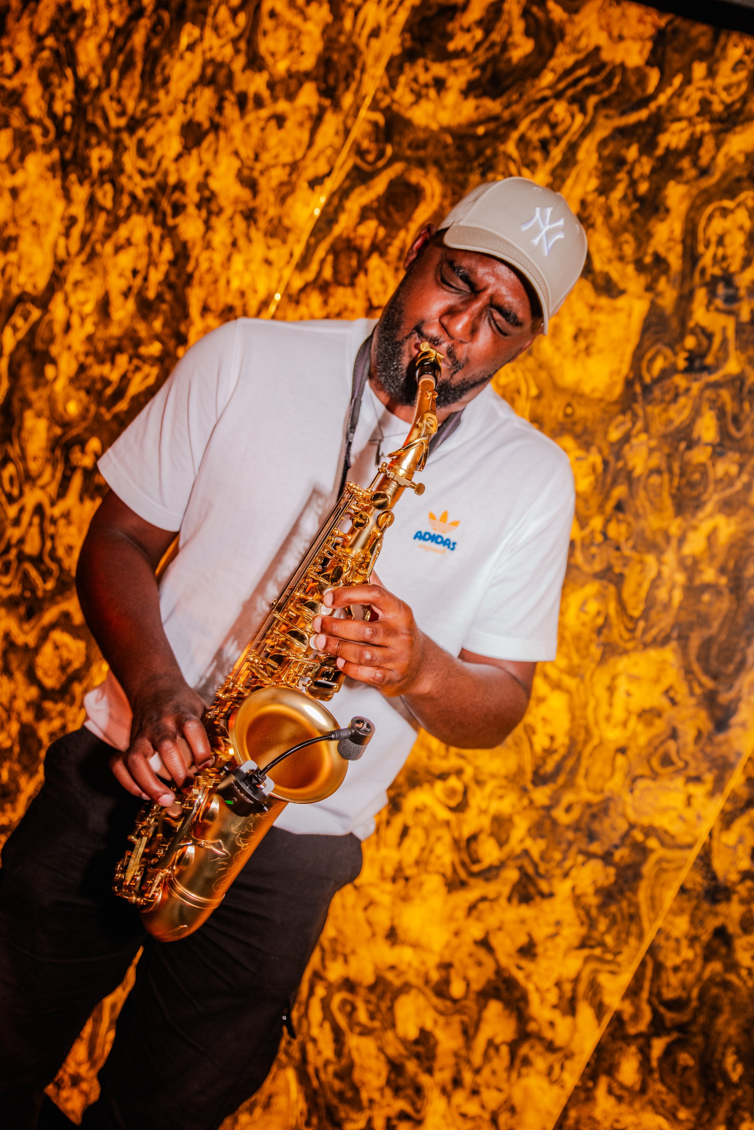 A man playing a gold saxophone, wearing a beige Yankees cap and a white Adidas shirt, with a decorative yellow and black patterned backdrop.