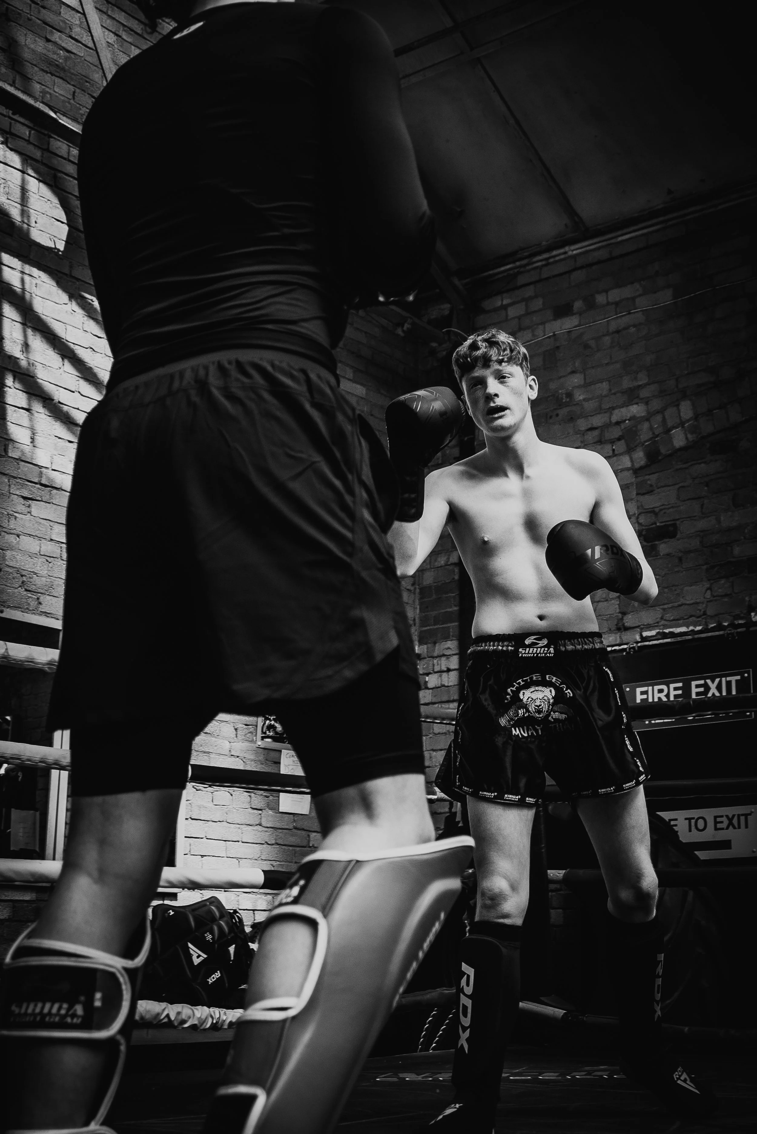 Two young men practicing boxing in a gym with brick walls, one is shirtless and wearing boxing gloves, the other is wearing a dark tank top and shorts, both are in a fighting stance inside a boxing ring.