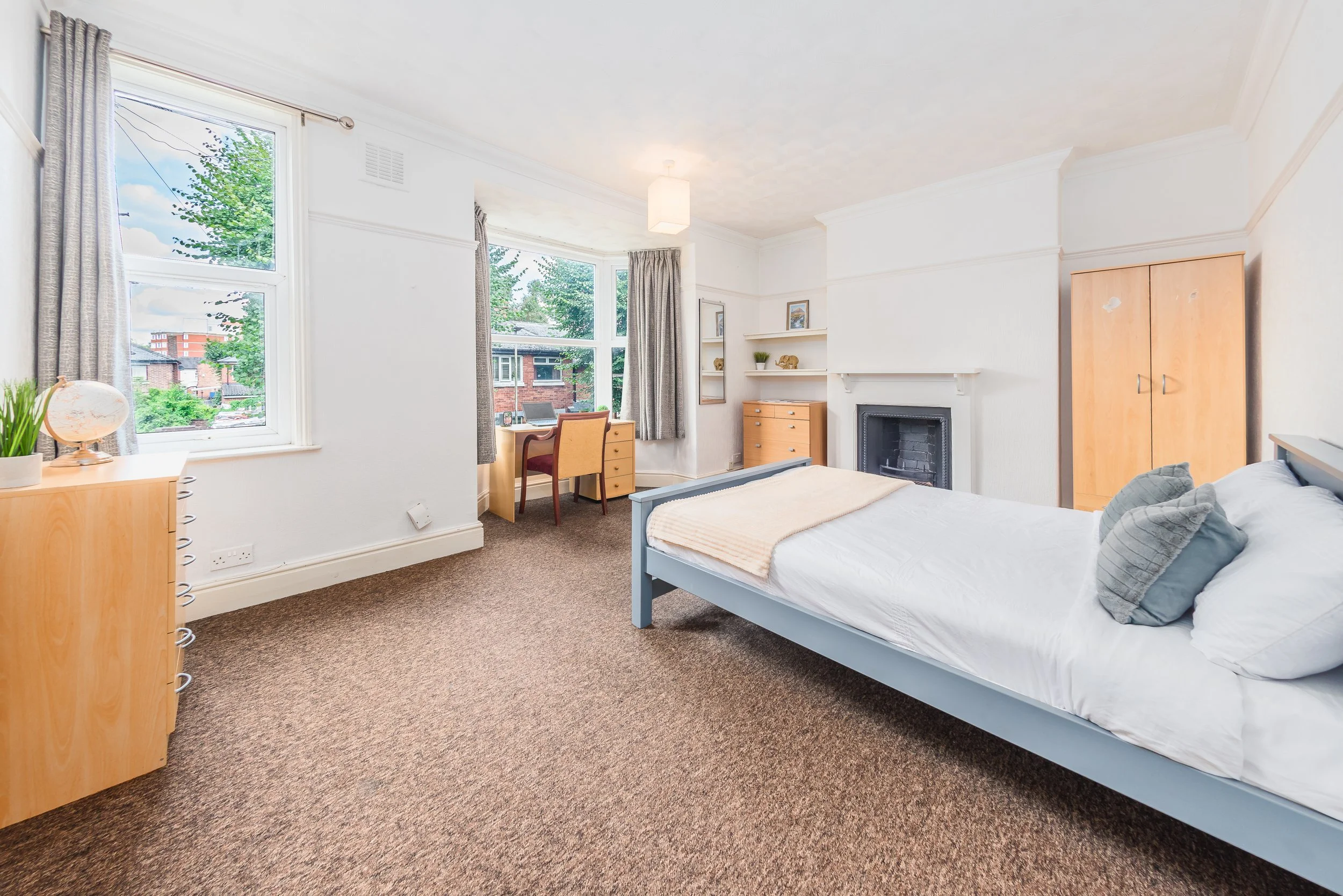 A bright bedroom with large windows, a bed with white sheets and pillows, wooden furniture including a wardrobe, desk, and chest of drawers, a fireplace, and decorative items on shelves.