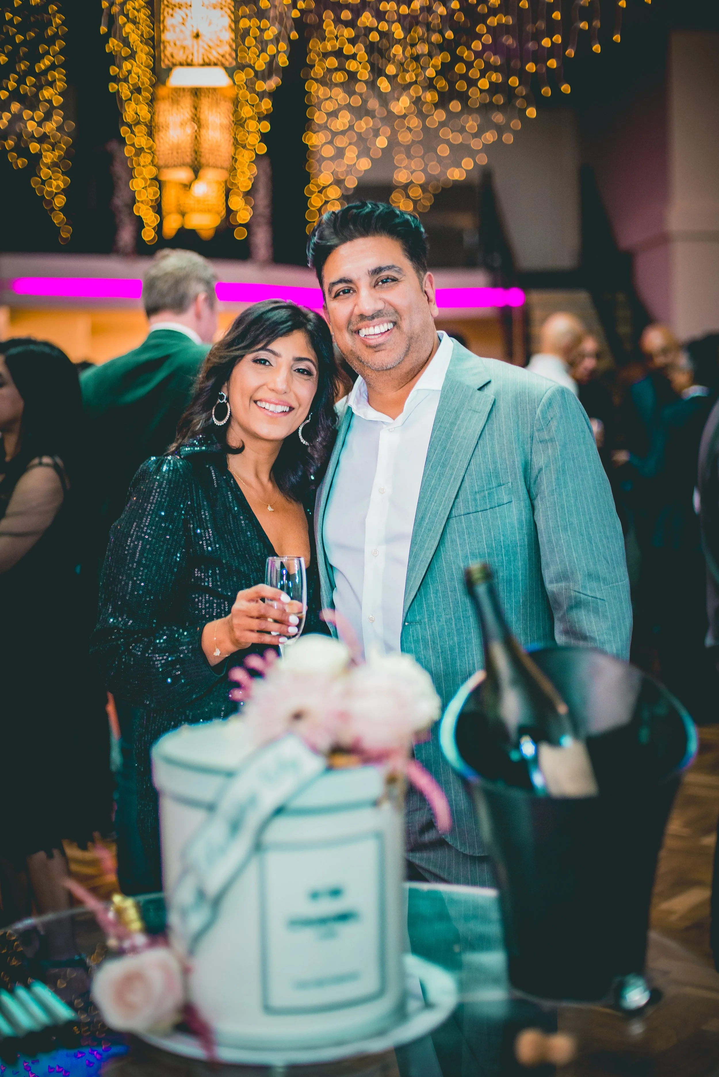 A smiling man and woman at a celebration, holding glasses of champagne, in a decorated event space with fairy lights and other guests in the background.