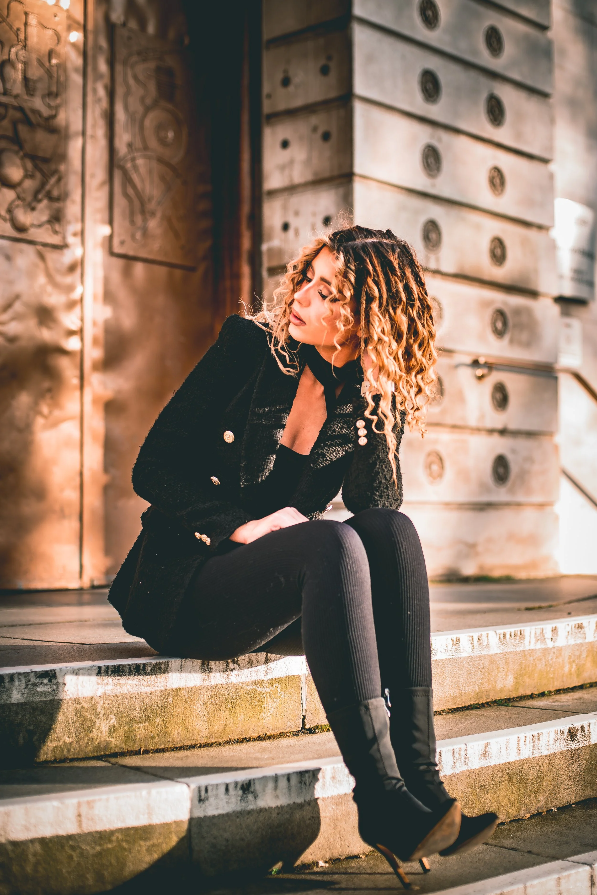 A woman with blonde, curly hair sitting on a concrete step outside. She is wearing a black jacket, black tights, and black boots. She has her head slightly tilted with her eyes closed, and her hair falls over her face. In the background, there are la