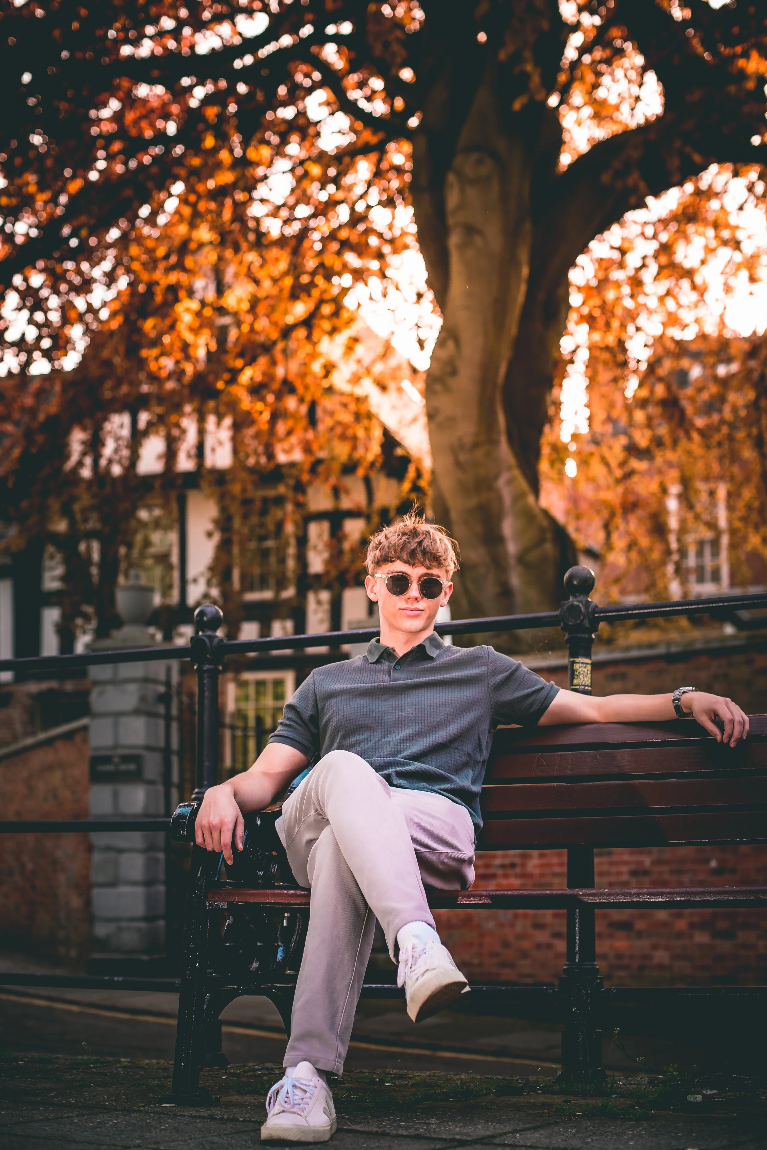 A man with curly hair, sunglasses, a dark gray polo shirt, and light-colored pants sitting on a park bench with his right leg crossed over his left, in front of a large tree with orange and red autumn leaves. The background includes buildings and a f