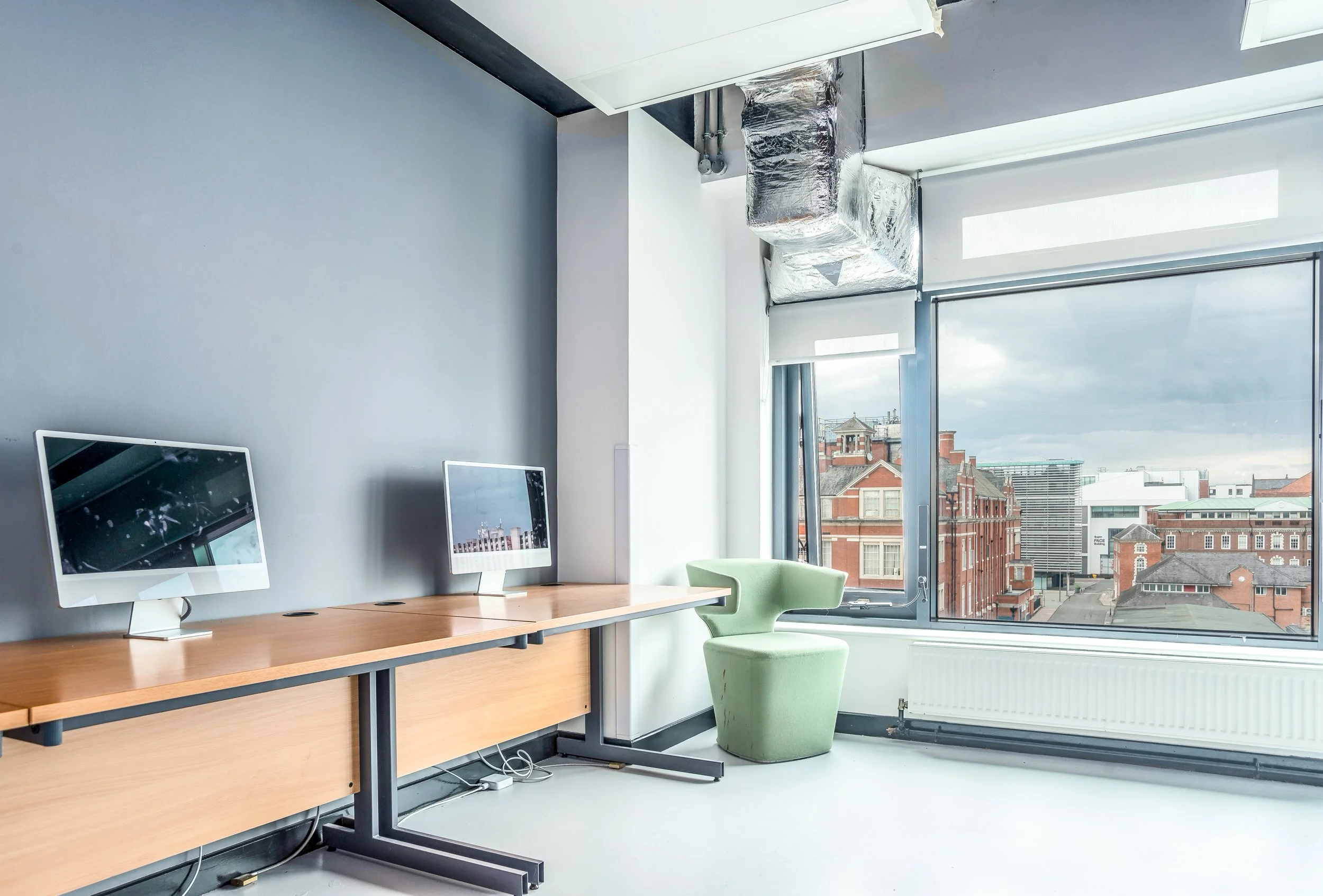 An office space with a wooden desk equipped with two desktop computers, a green ergonomic chair, and large windows showing a cityscape with buildings under a cloudy sky.
