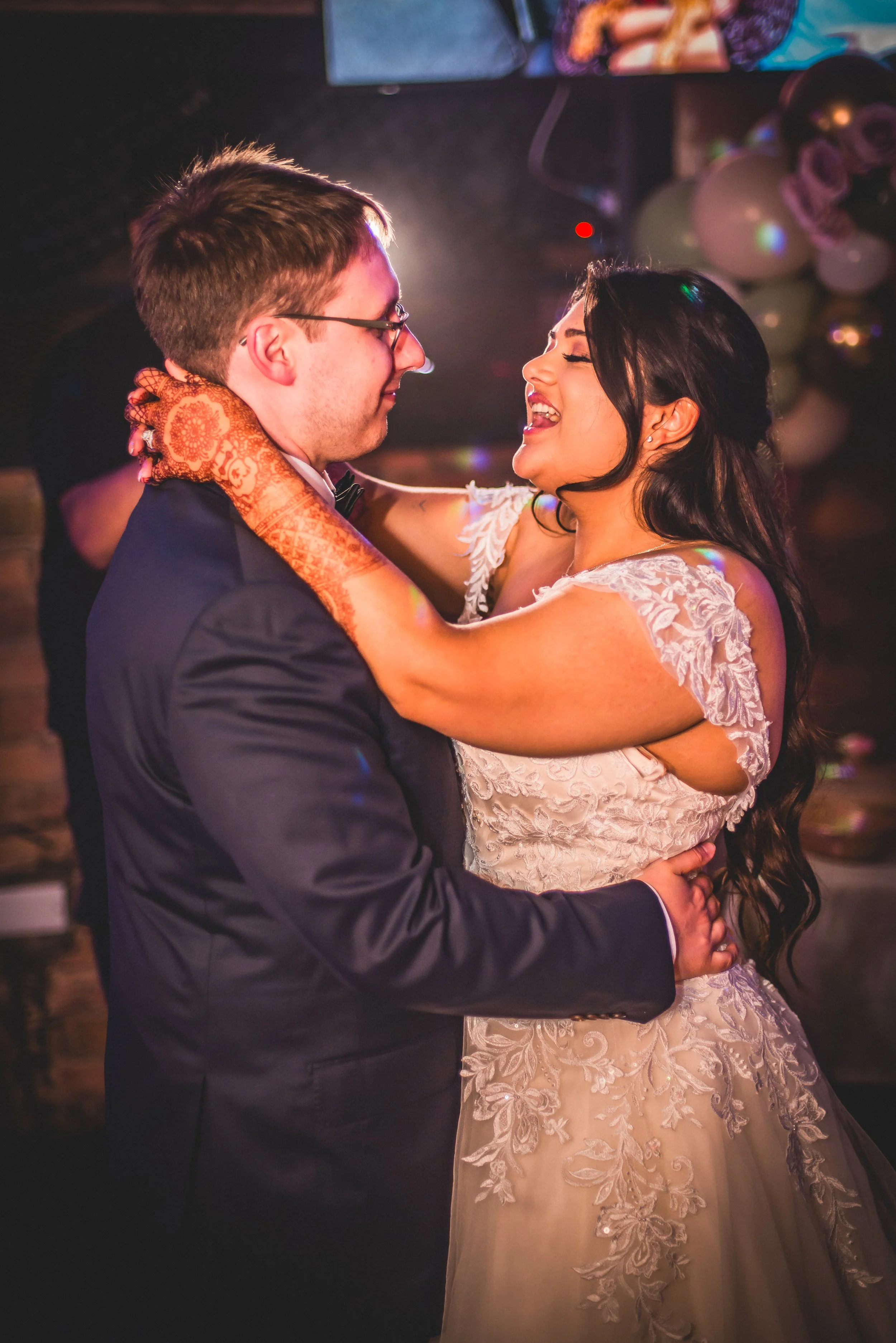 A couple dancing closely at a wedding reception, with the woman in a lace wedding dress and the man in a tuxedo. They are smiling and looking at each other.
