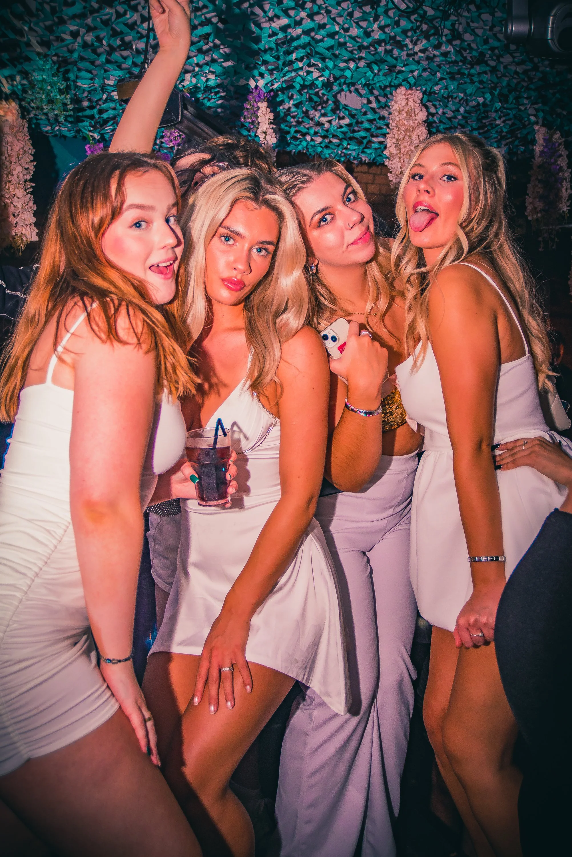Group of five young women dressed in white at a party, posing closely together, some with their tongues out, one holding a drink, in a decorated venue with hanging flowers and colorful decorations.