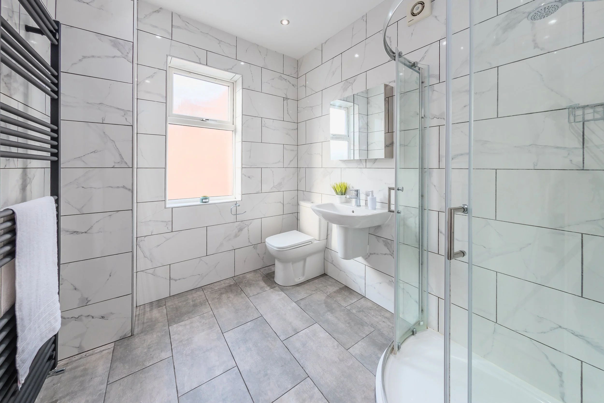 A modern bathroom with white marble tiled walls, a small window, a toilet, a pedestal sink with a mirror, a potted plant, a glass shower enclosure, and a black heated towel rail with white towels.
