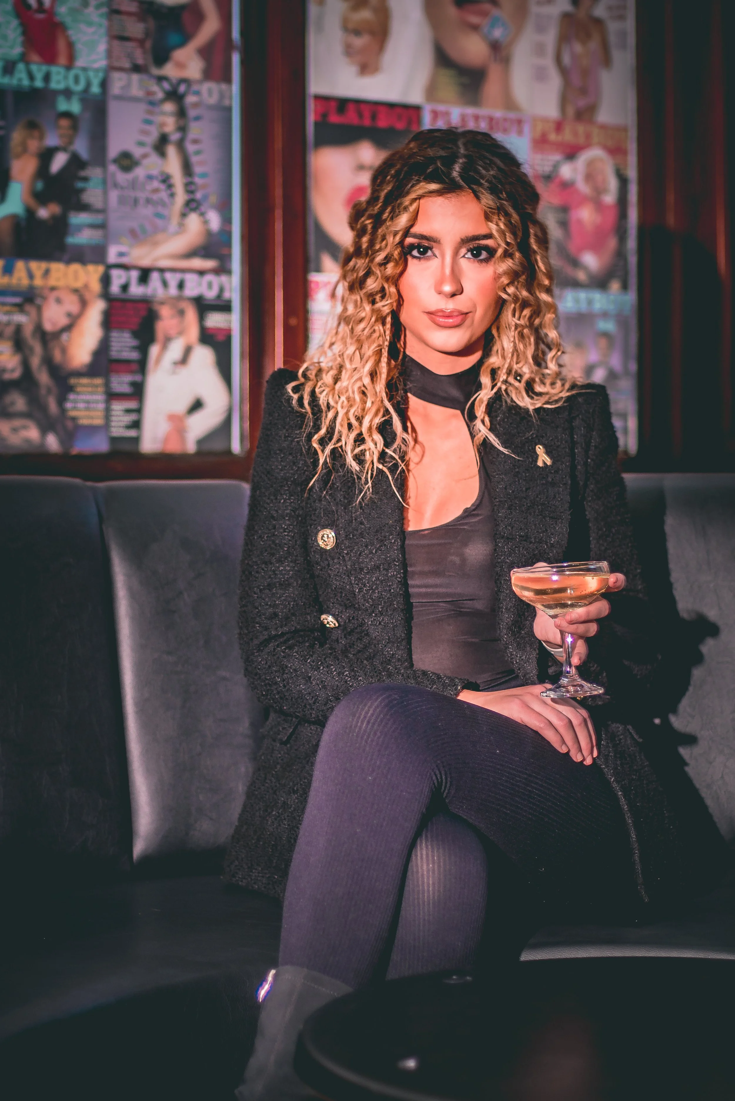 A woman with curly blonde hair and makeup, wearing a black blazer with gold buttons, sitting on a black leather sofa in a room decorated with Playboy magazine covers.