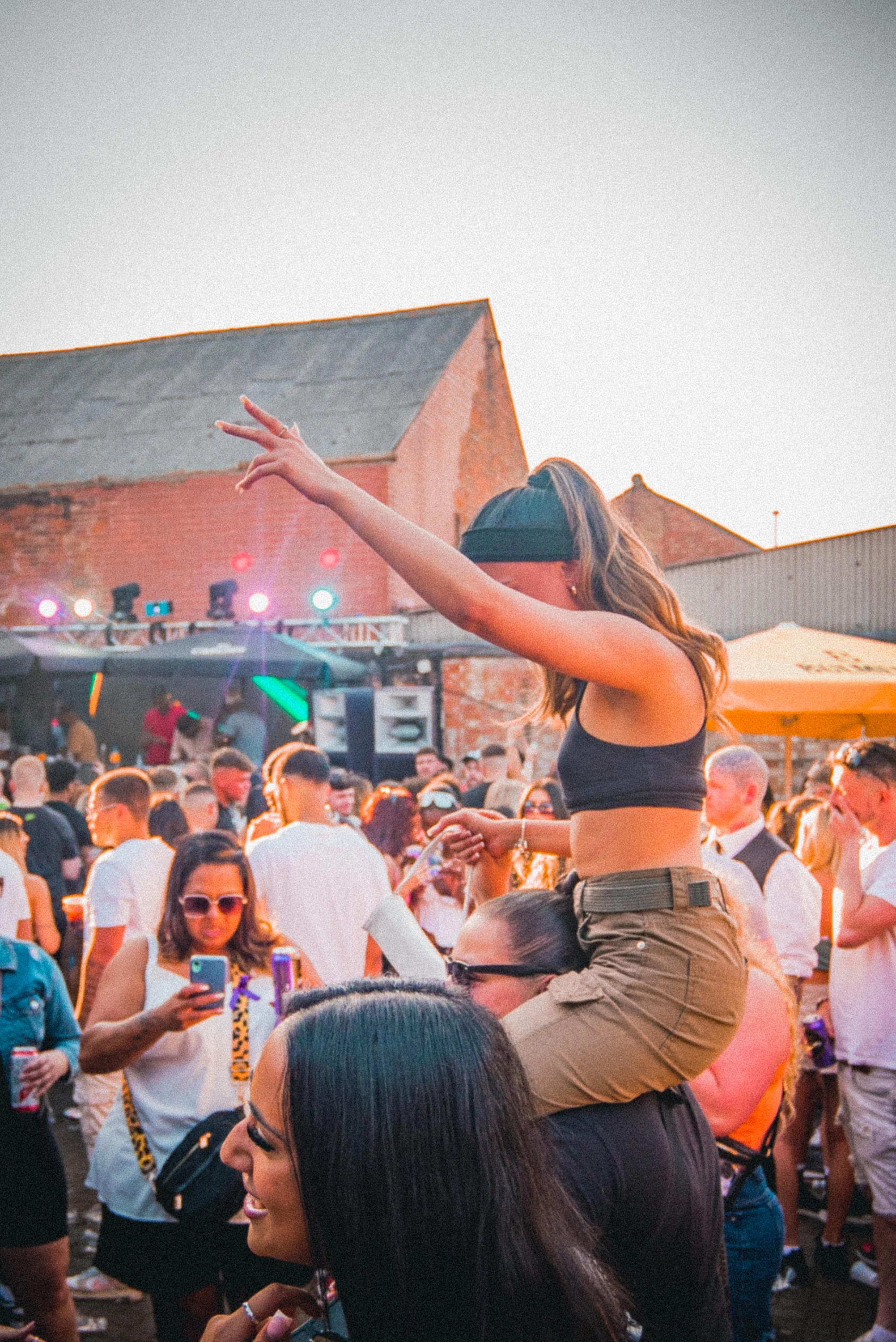 A young woman with sunglasses and long hair, making a peace sign with her hand while standing on someone's shoulders at a crowded outdoor event during sunset.
