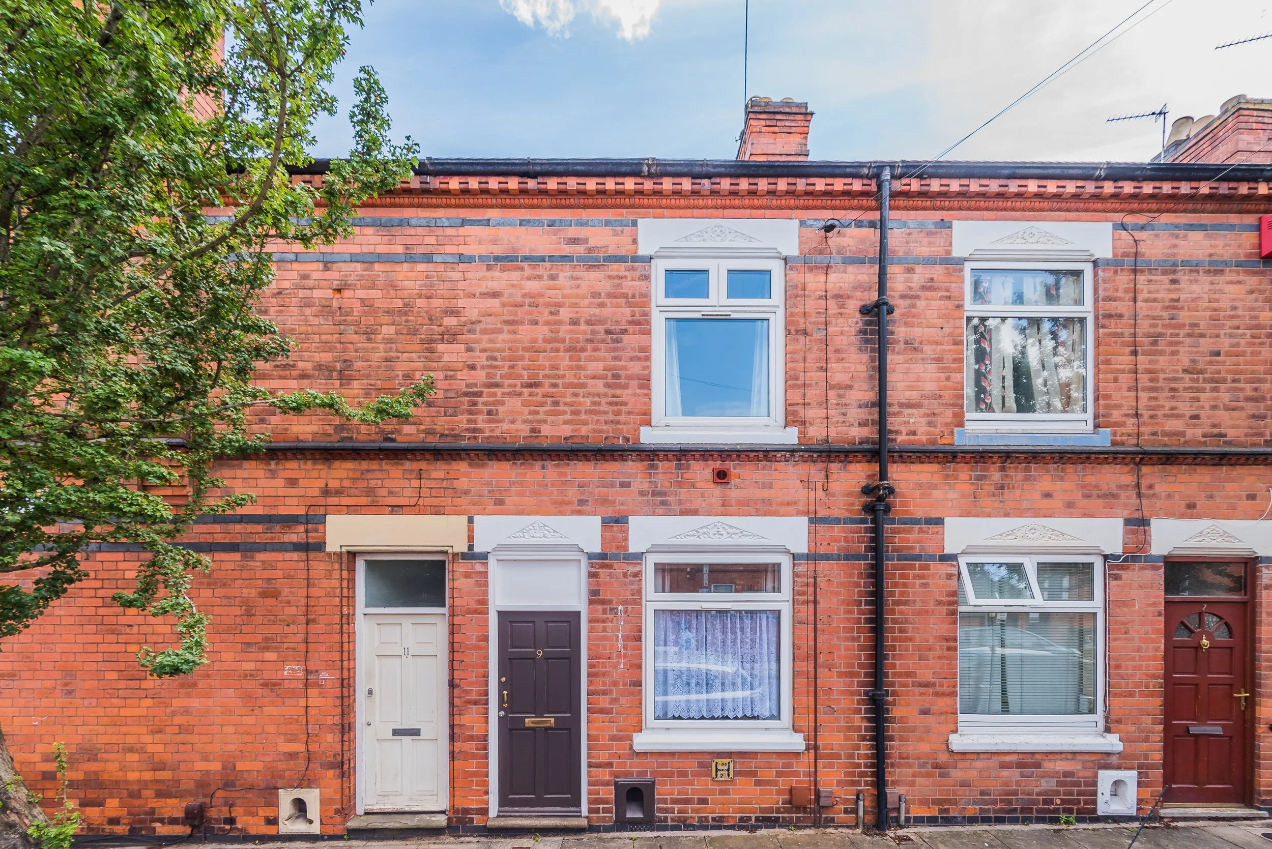 A row of red brick townhouses with white and brown doors, a large window, and a smaller window. A tree with green leaves is visible on the left side of the image. The sky is partly cloudy.