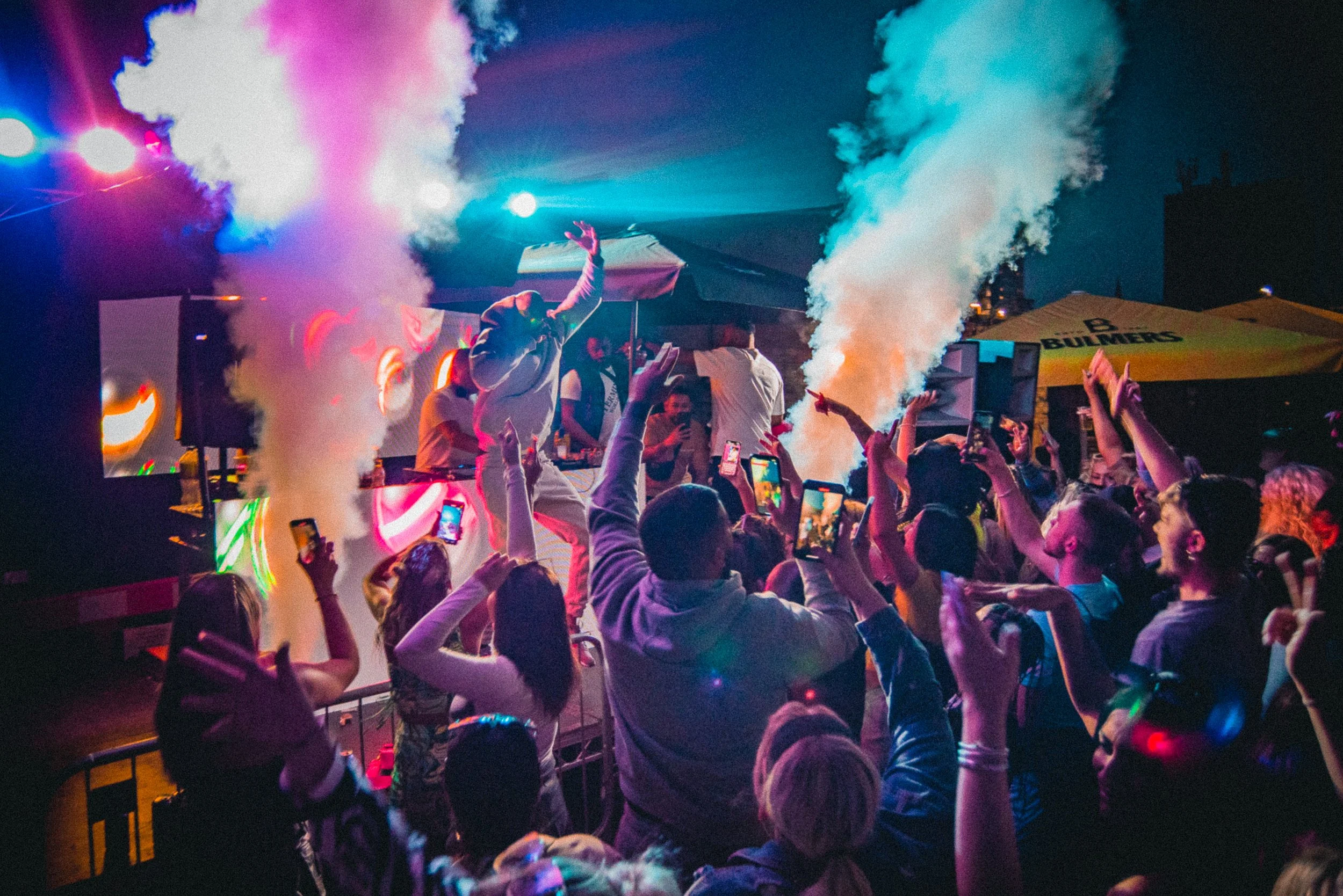 A large crowd of people at a nighttime outdoor concert or festival, with colorful lighting and smoke effects, some raising their hands and taking photos.