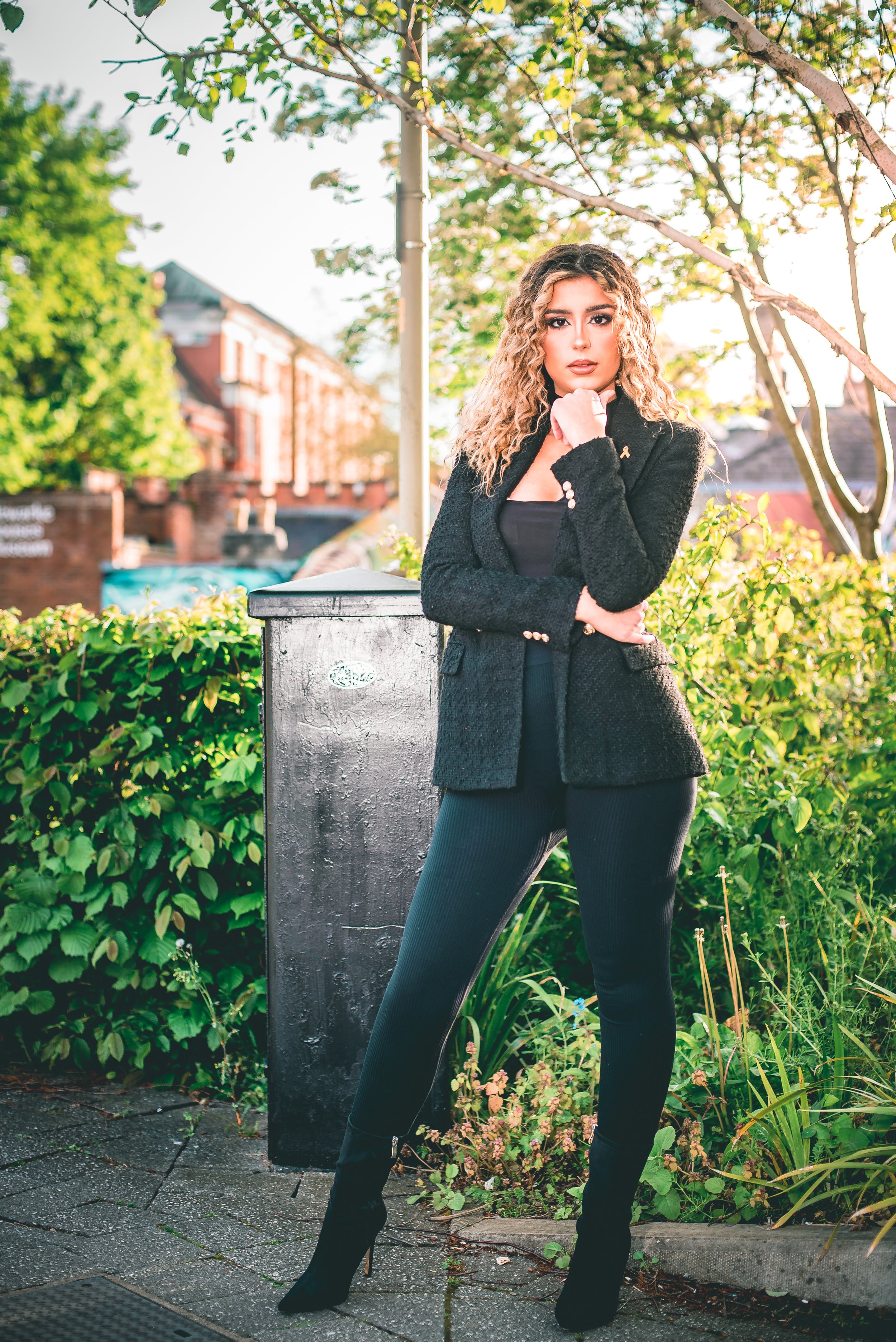 A woman with curly blonde hair poses outdoors, dressed in a black blazer and pants, standing next to a black utility box, with greenery and a brick building in the background.