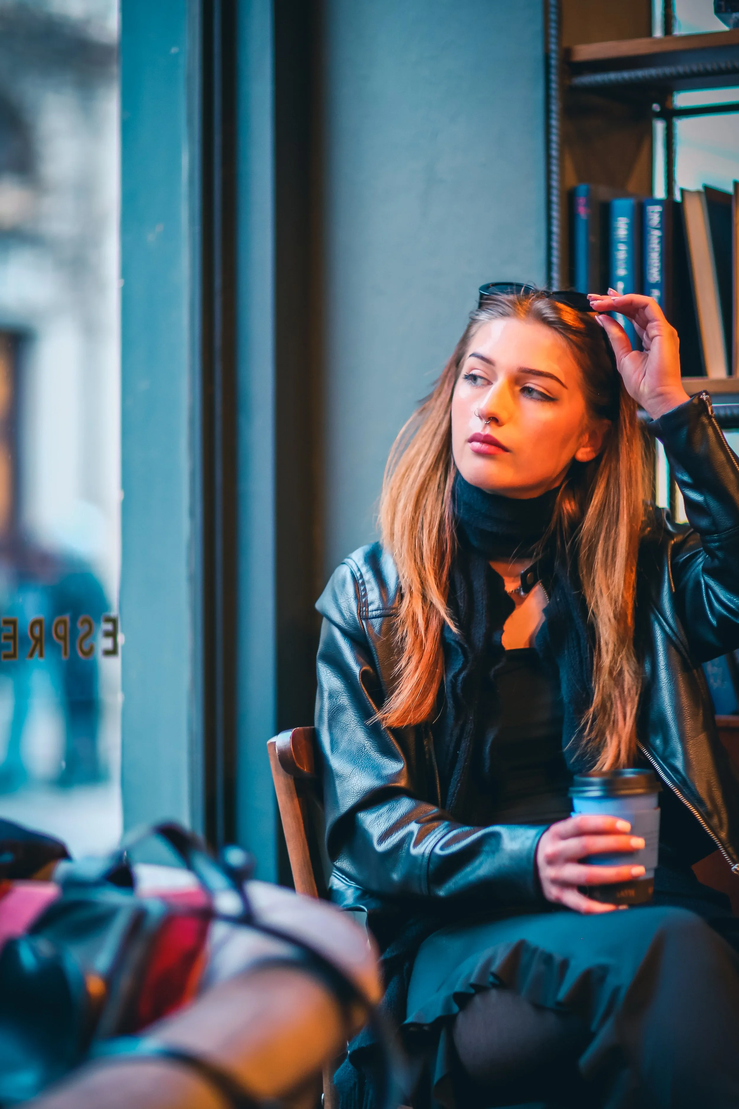 A young woman with long hair, wearing a black turtleneck and a leather jacket, sitting in a coffee shop, holding a paper coffee cup, with shelves of books in the background.