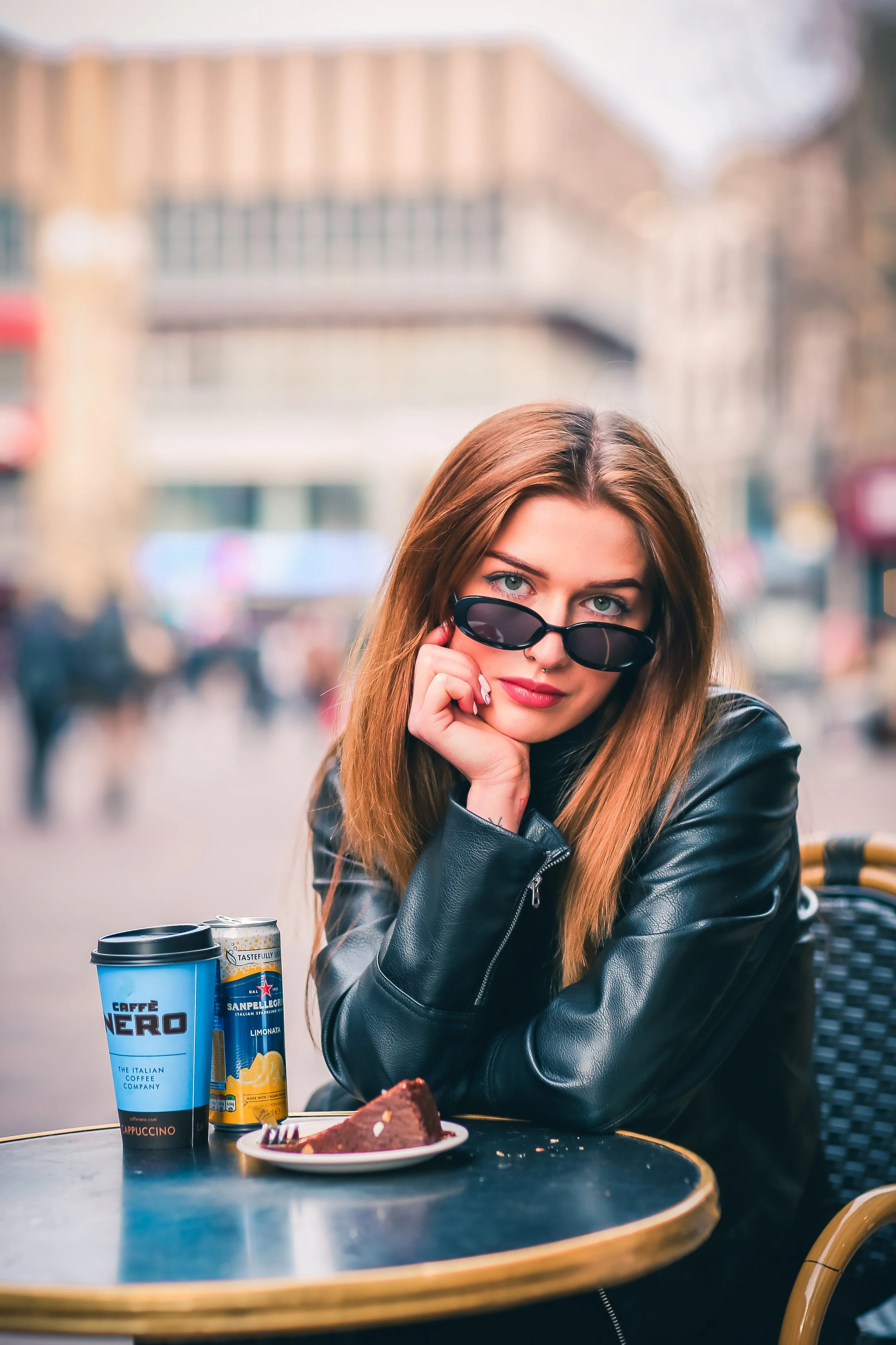 Young woman with long brown hair and blue eyes, wearing a black leather jacket, is looking over her sunglasses at a café table with a slice of chocolate cake, two cans of San Pellegrino sparkling water, and a blue and black coffee cup.