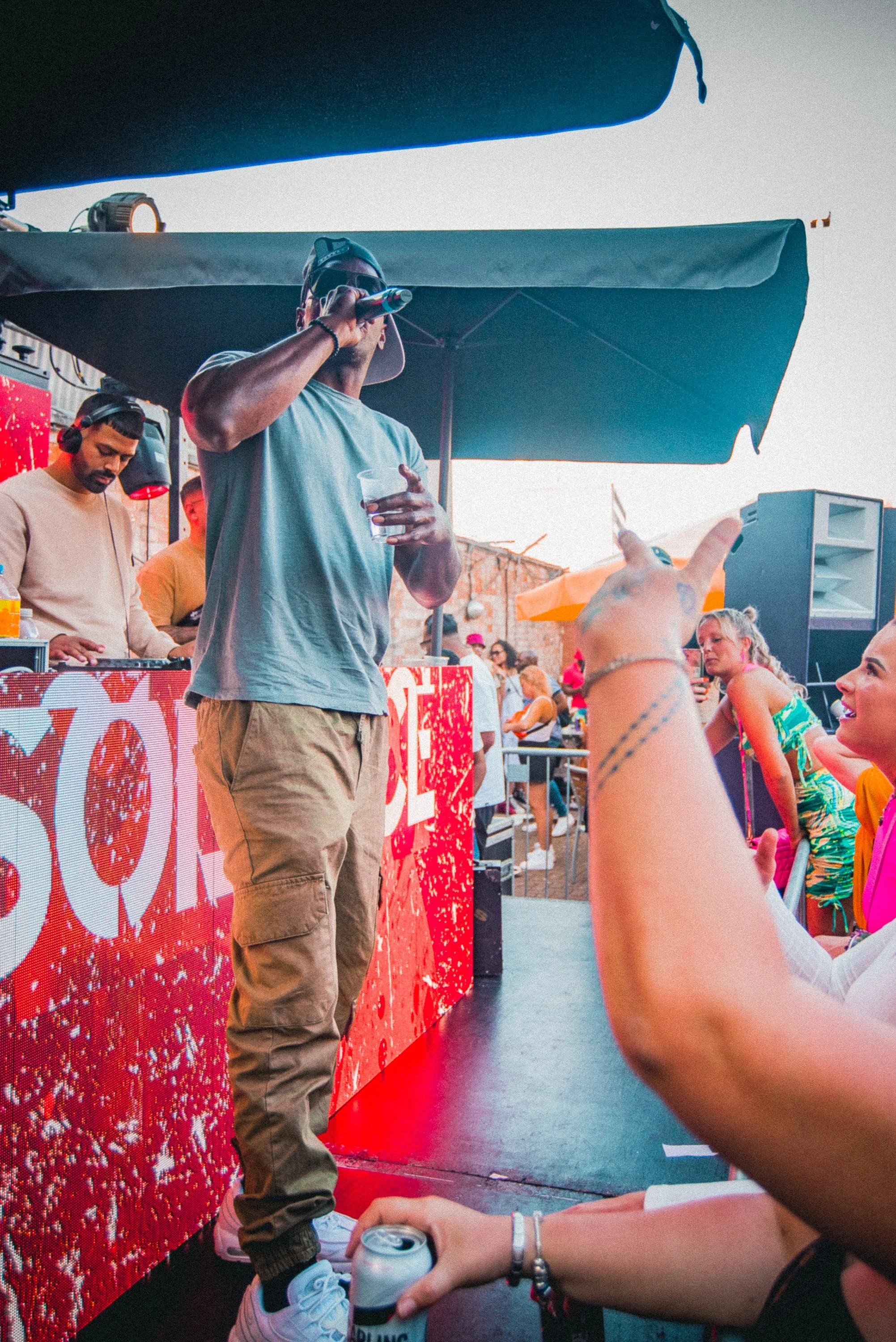 A man stands behind a bar at an outdoor event, speaking into a microphone. He holds a clear cup with a drink in one hand and wears sunglasses and a backward baseball cap. Several people are in the background, and one person in the foreground is handi