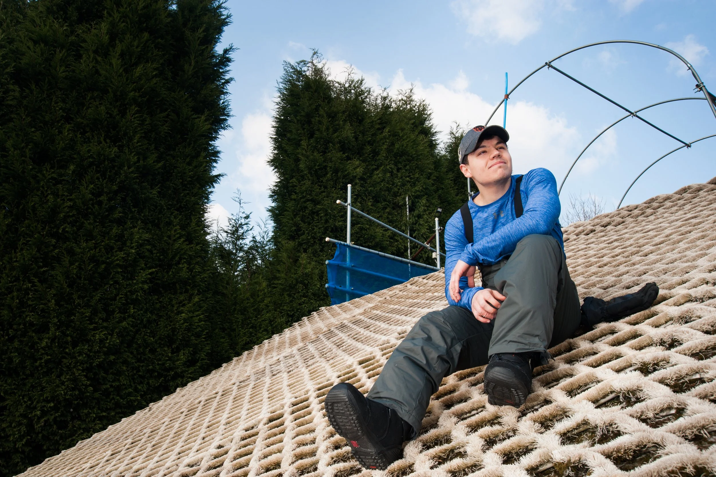 A man sitting on a rooftop made of shingles, wearing outdoor clothing and a cap, with tall evergreen trees and a partly cloudy sky in the background.