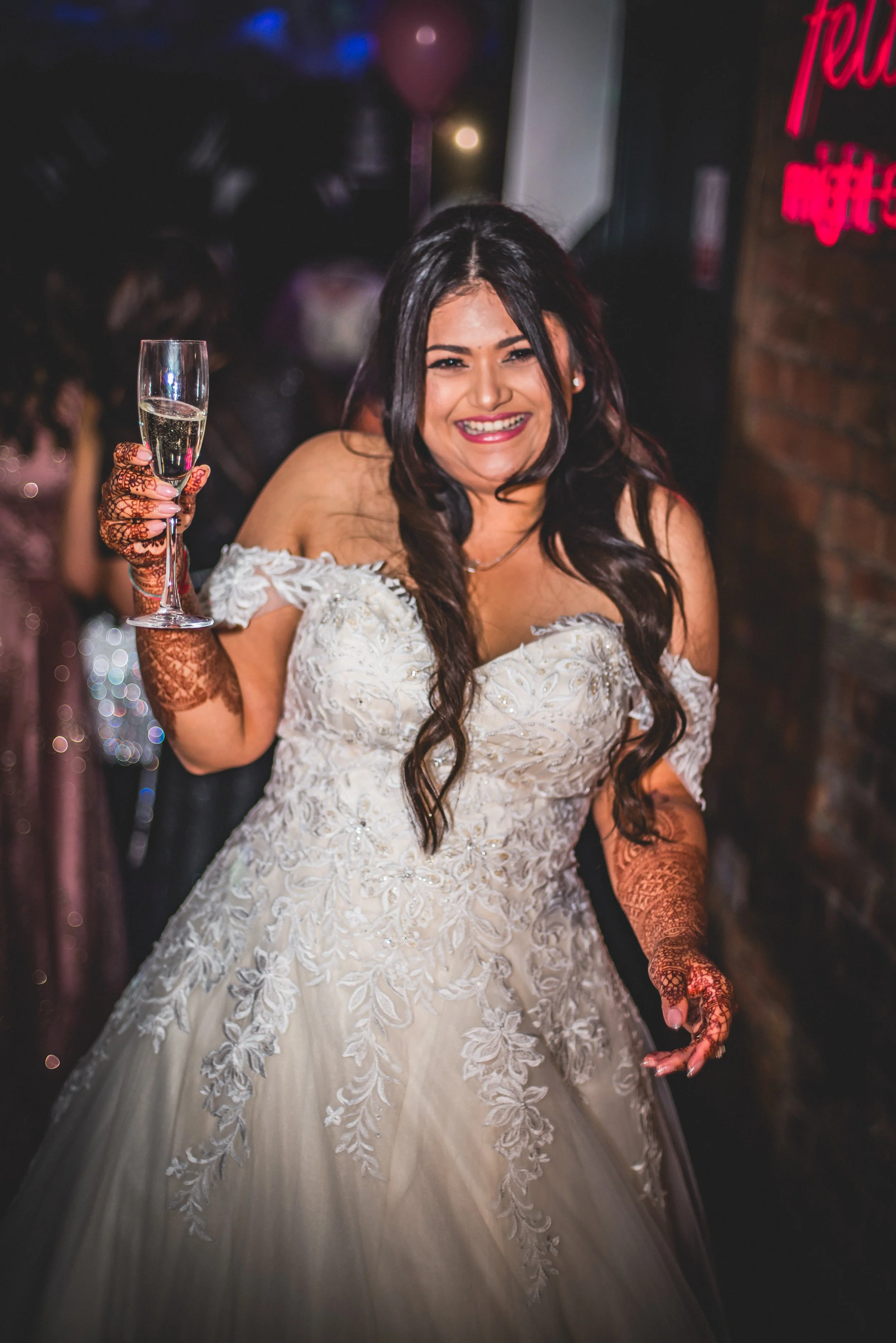 Woman in an off-shoulder white wedding dress smiling, holding a glass of champagne. She has long dark hair and intricate henna designs on her hands and arms, in a dimly lit venue with dark walls and neon signs.
