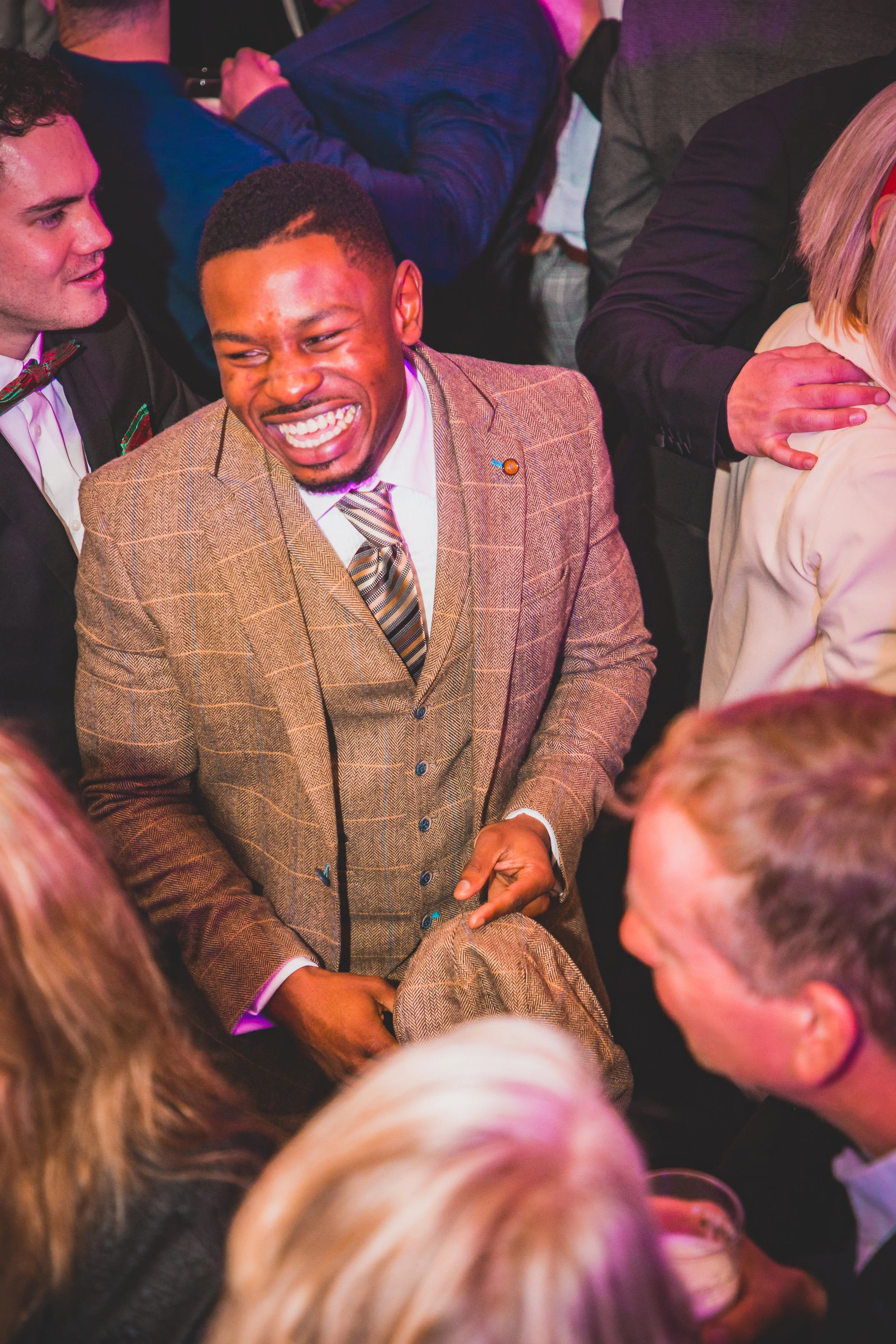 A group of people at a social gathering, with one man in the center wearing a brown plaid suit laughing, surrounded by others engaged in conversations.