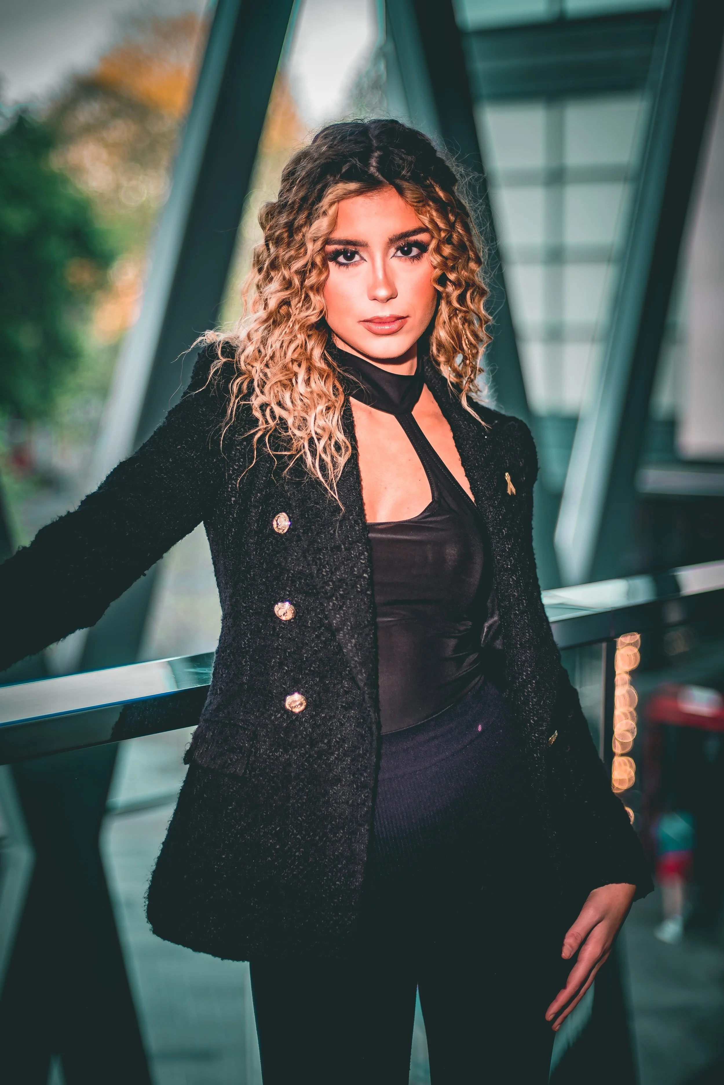 A woman with curly blonde hair and striking makeup, wearing a black outfit and a textured black blazer, standing in front of a modern architectural background with large windows and green trees outside.