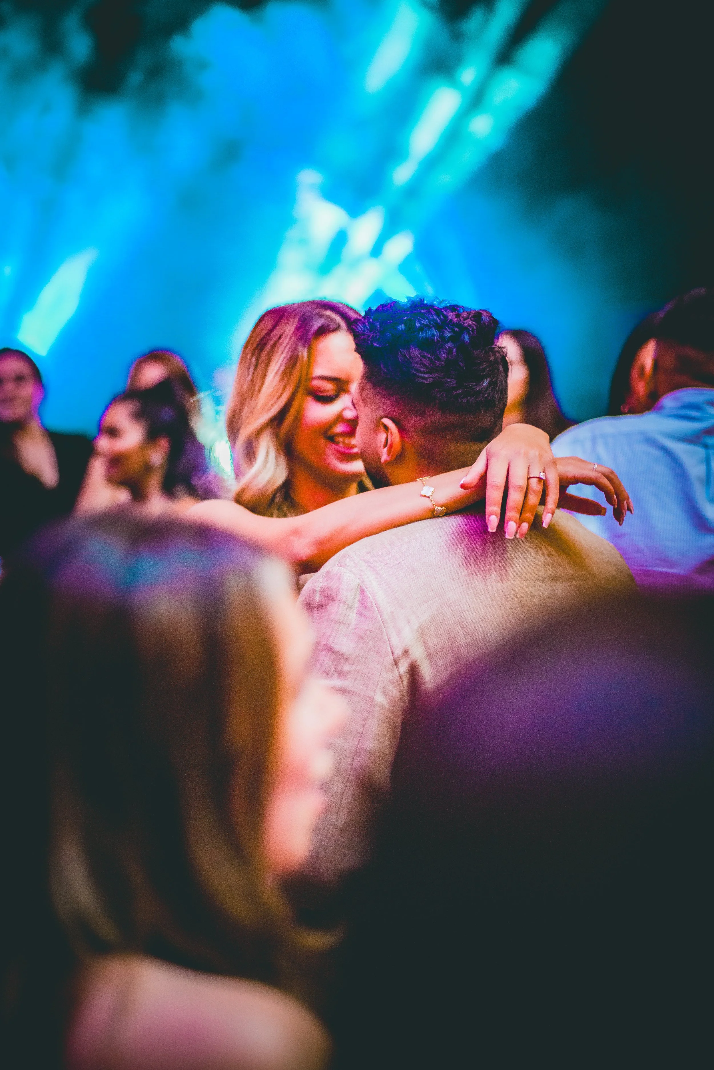 A couple dancing closely at a lively party or nightclub with blue and purple lighting, surrounded by other people.