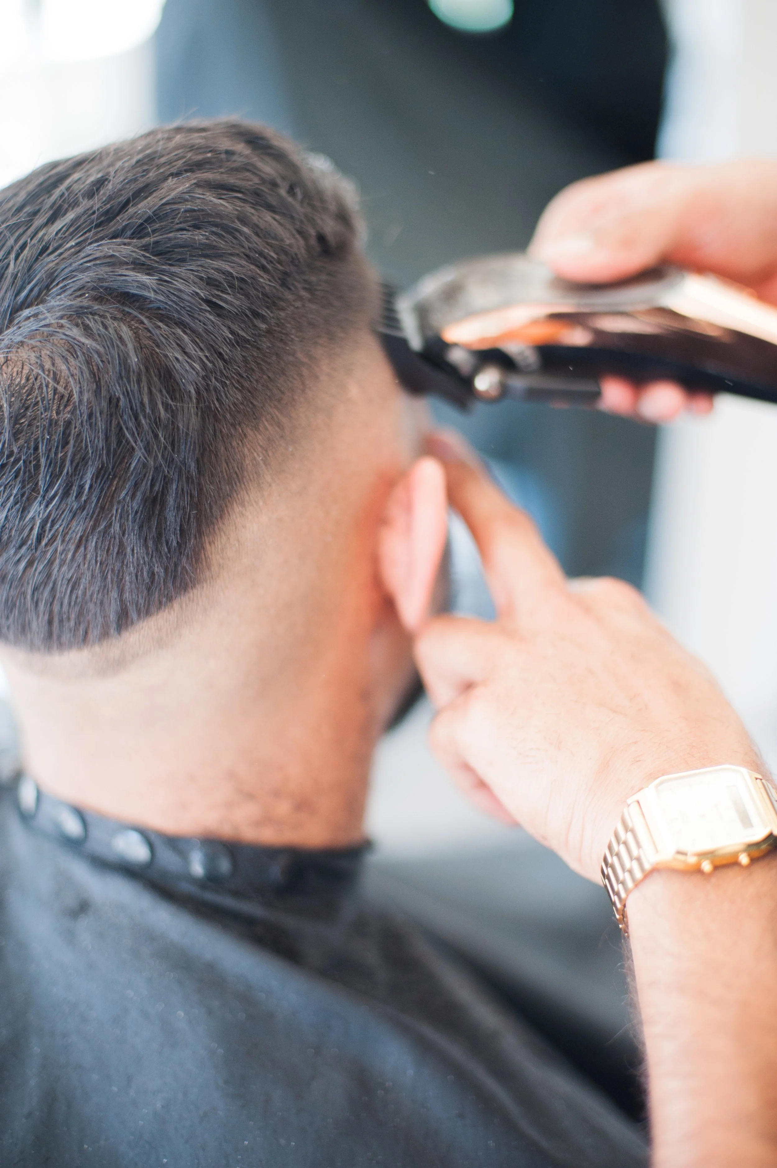 Close-up of a person having their hair cut with hair clippers at a salon or barbershop, wearing a black cape and a gold wristwatch.