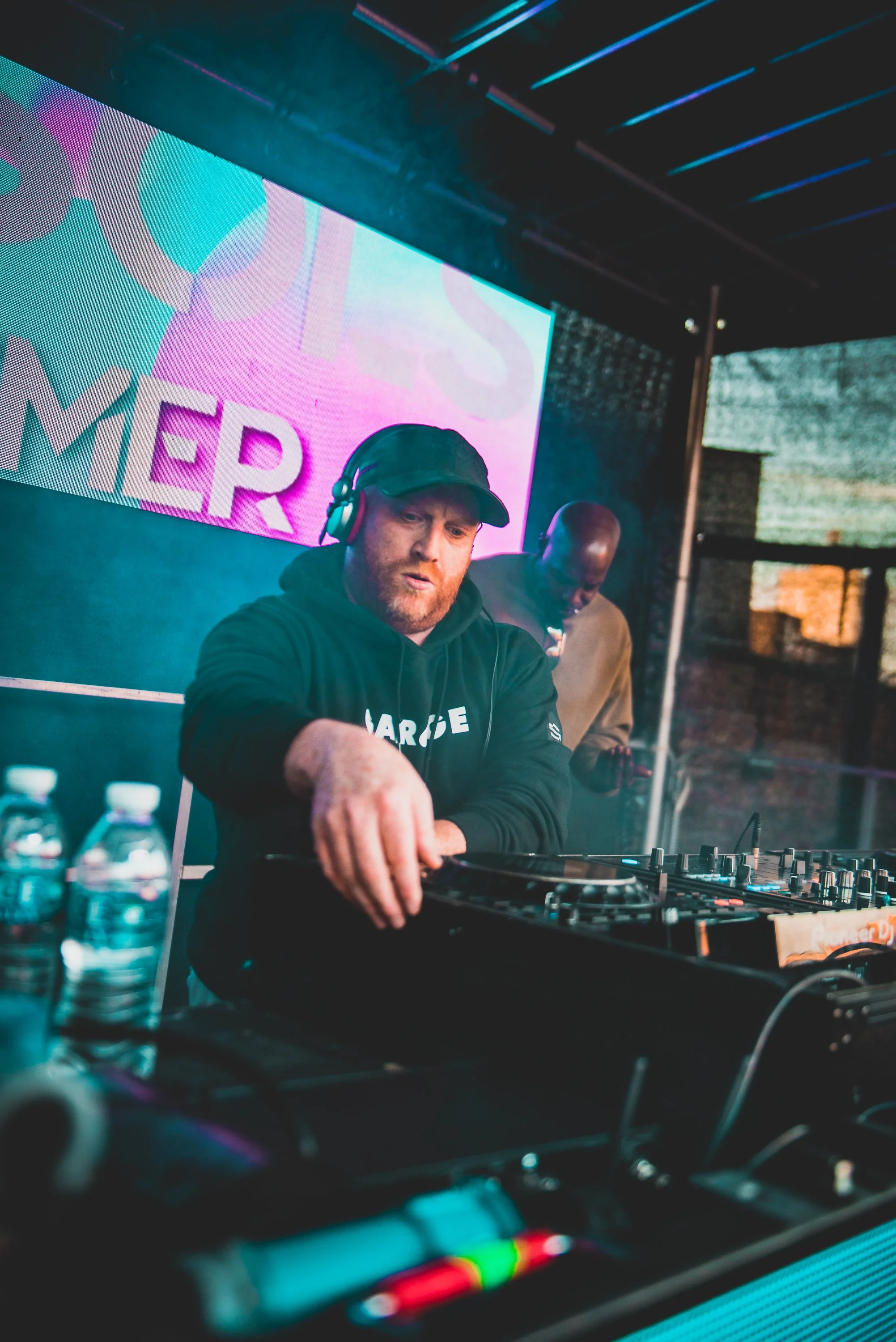 A DJ with a beard, wearing a black hoodie and a cap, is performing at a DJ booth with turntables and equipment. Behind him is a large screen displaying colorful graphics and the word 'HUIS'.
