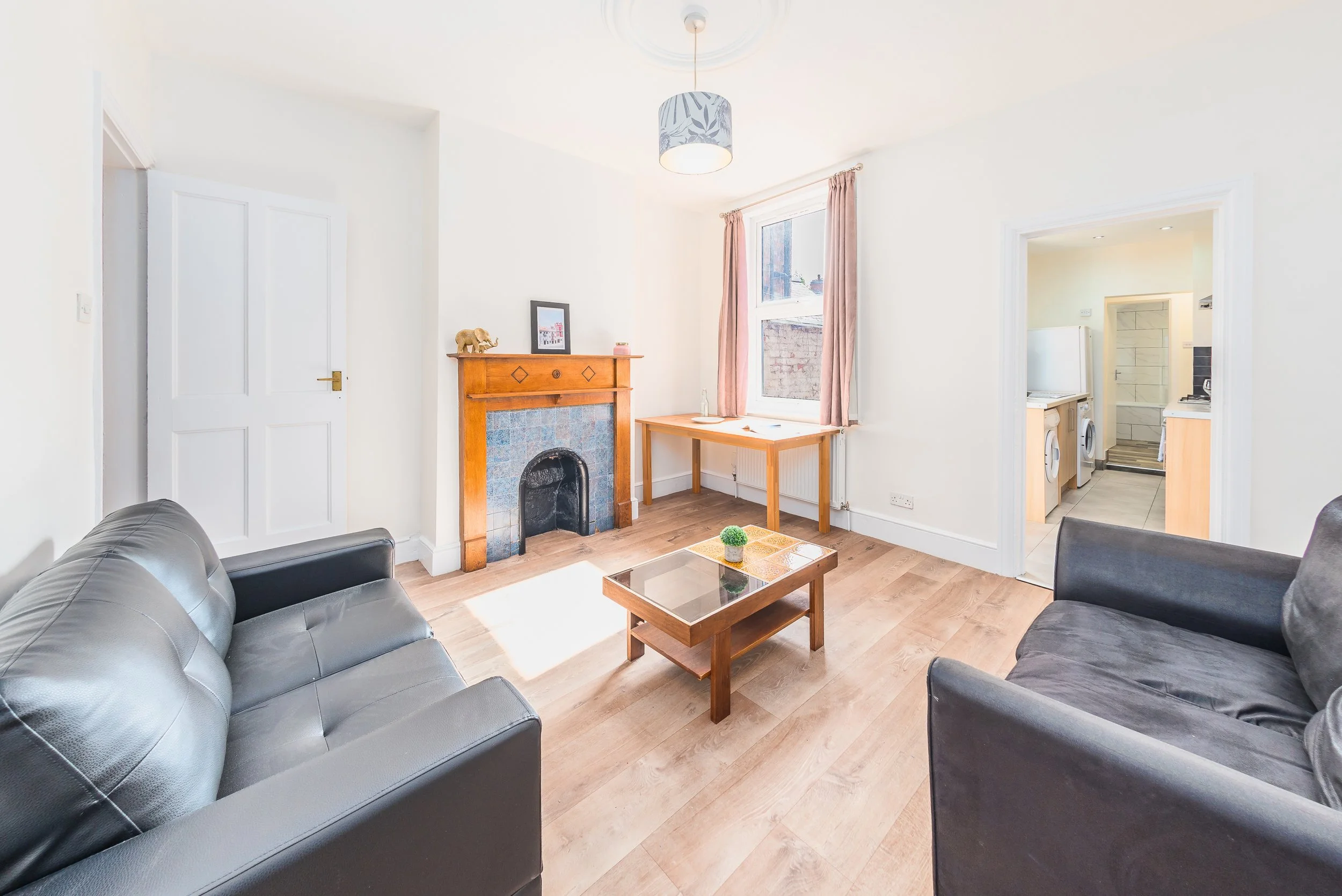 Living room with two black leather couches, wooden coffee table, fireplace with decorative items, large window with pink curtains, view into kitchen with washing machine and stove.