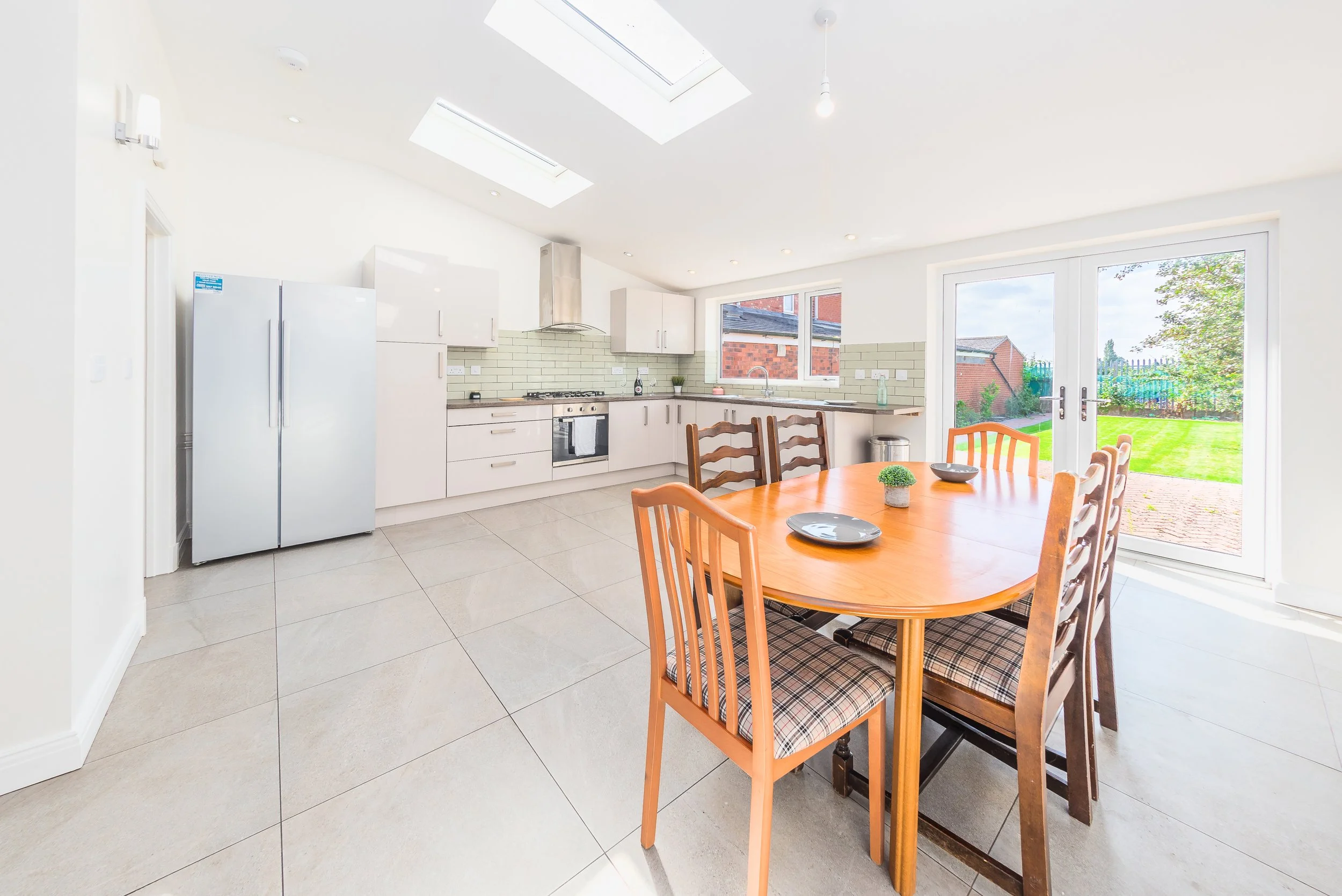 Bright, modern kitchen with white cabinetry, stainless steel oven, fridge, and range hood. Green tile backsplash, large window, sliding glass door overlooking a backyard with grass, trees, and a fence. Wooden dining table with matching chairs. Skylig