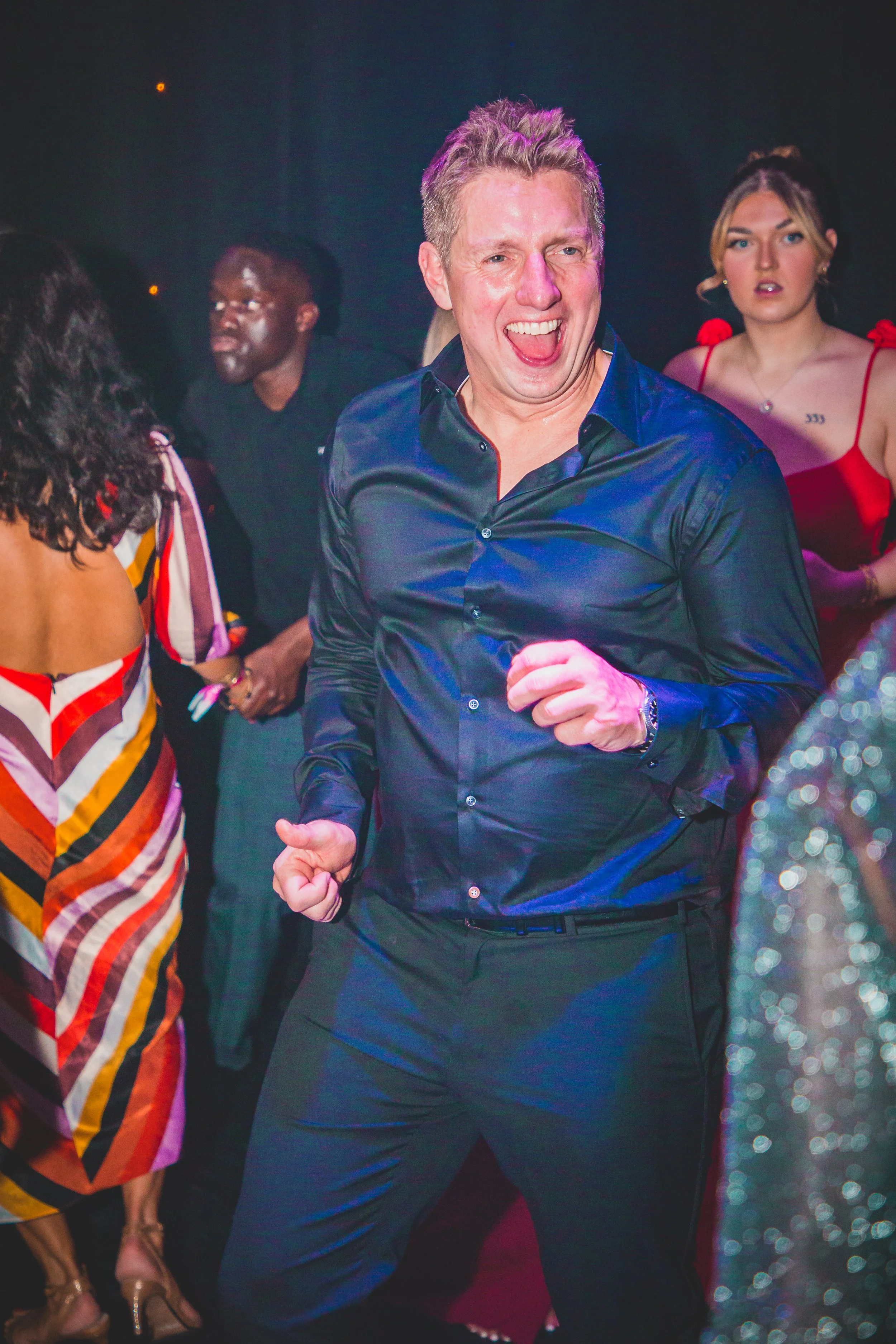 A man in a black dress shirt dancing and smiling at a party, surrounded by women in colorful dresses.
