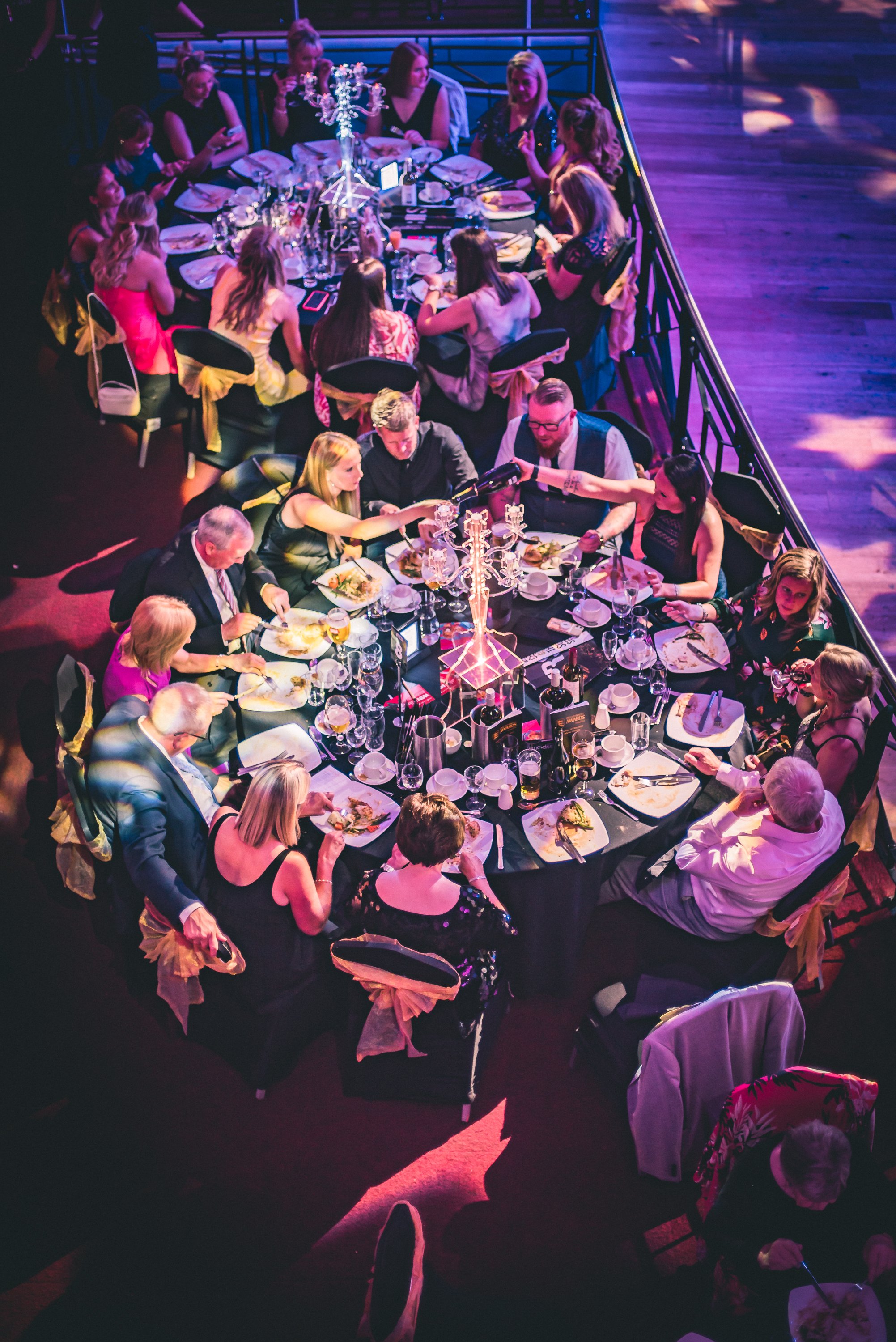 People seated around a large round dining table at a formal event, with elegant table settings, a decorative candelabra, and a dark wooden dance floor with colorful lighting in the background.