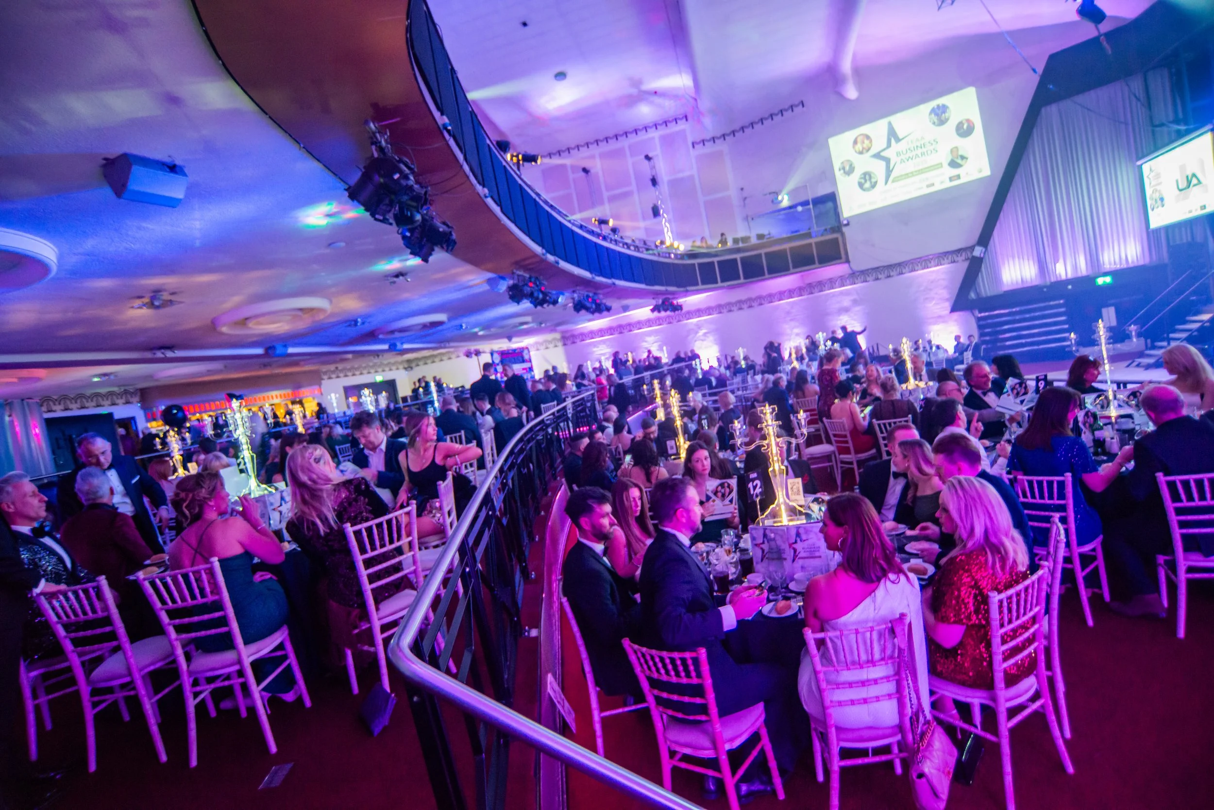 A large indoor event with round tables filled with people dressed in formal attire, some with glasses and suits, attending a ceremony or awards event. There are multiple screens displaying the text 'Business Awards' and a stage in the background, ill