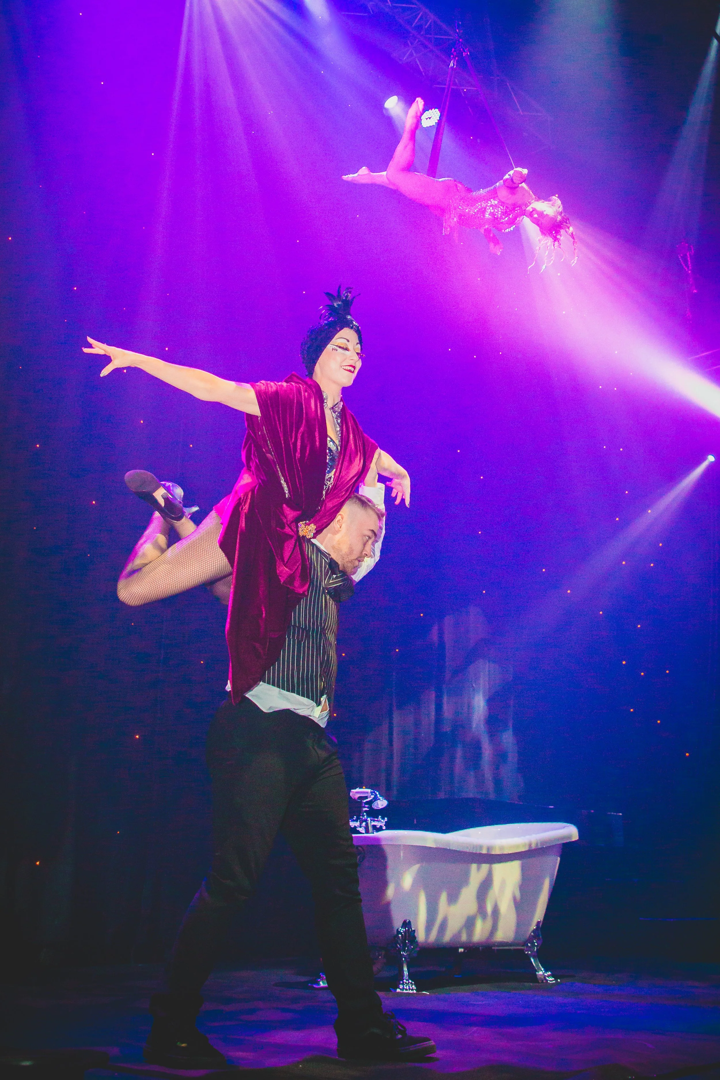 A circus performer dressed in a fancy costume with a top hat and coat holding another performer who is lying horizontally on their shoulders, with a purple and blue stage background and a prop resembling a bathtub.
