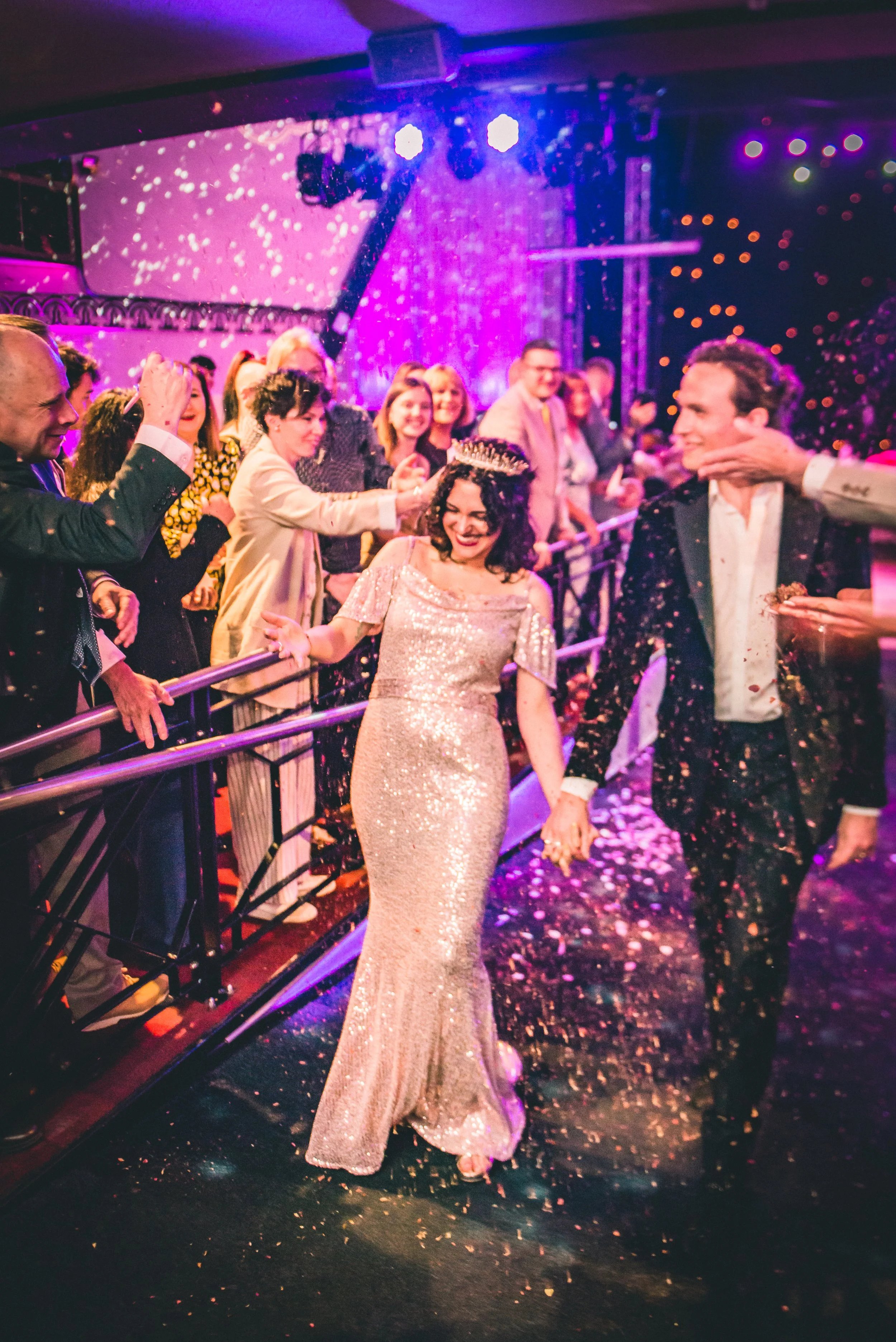 A woman in a sparkling floor-length gown and tiara smiling as she walks through a celebration with confetti, surrounded by people in formal attire at a lively party or event.