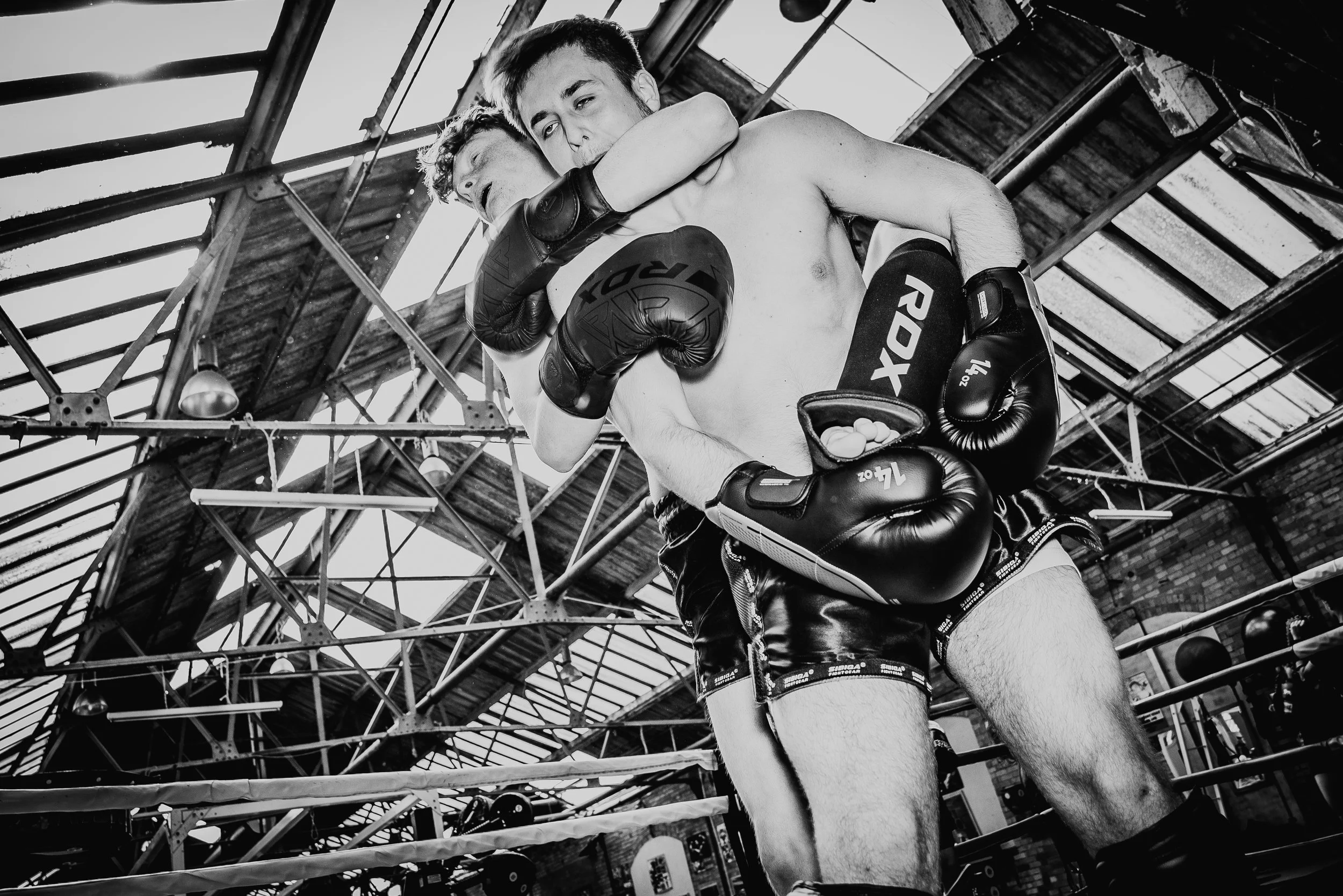 Two shirtless male fighters wearing gloves, embracing in a clinch inside a boxing ring with a wooden ceiling and scaffolding above.