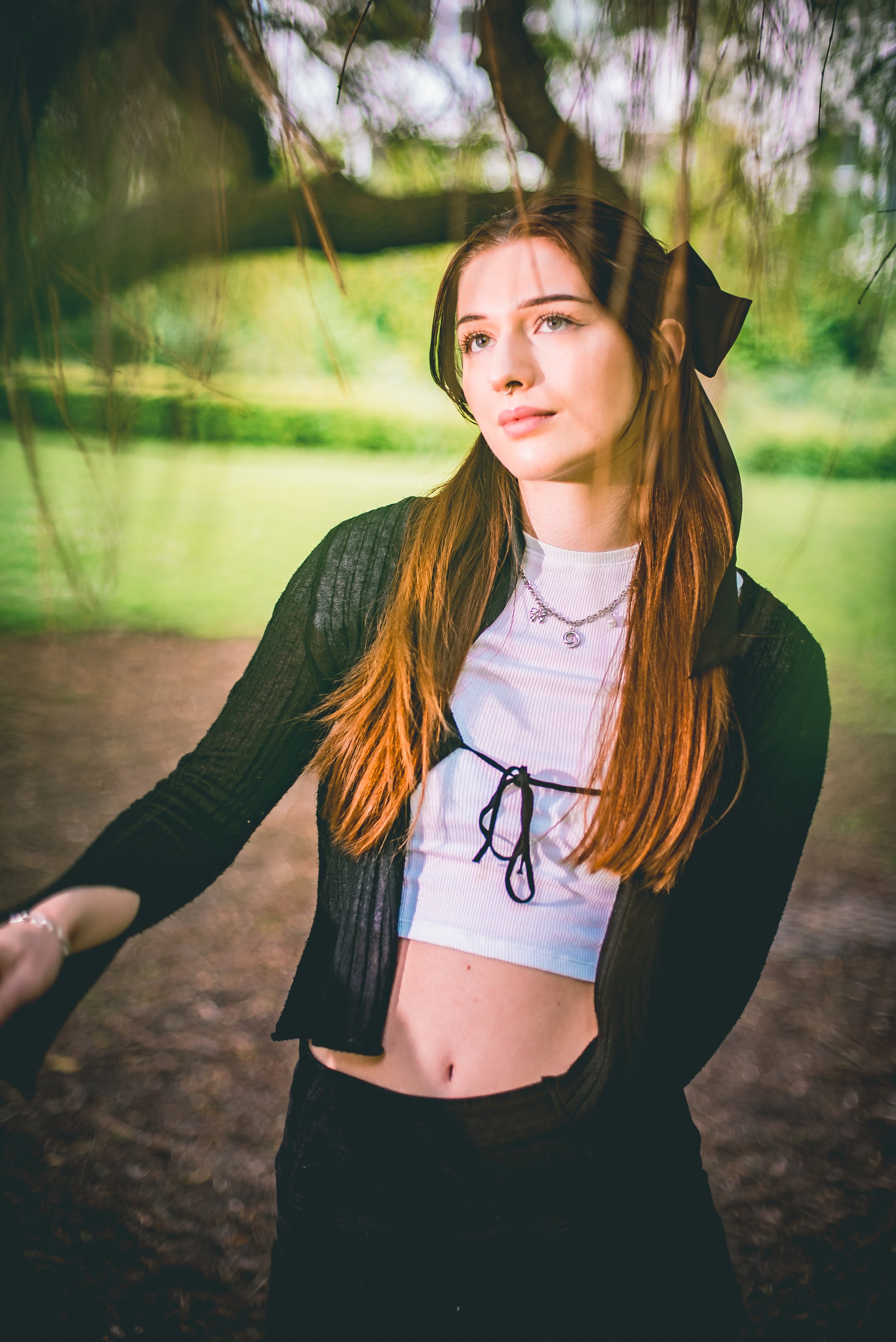 Young woman with long auburn hair standing outdoors, wearing a white crop top, black lace shorts, and a black sheer cardigan, with jewelry including necklaces and rings, under a tree in a park.