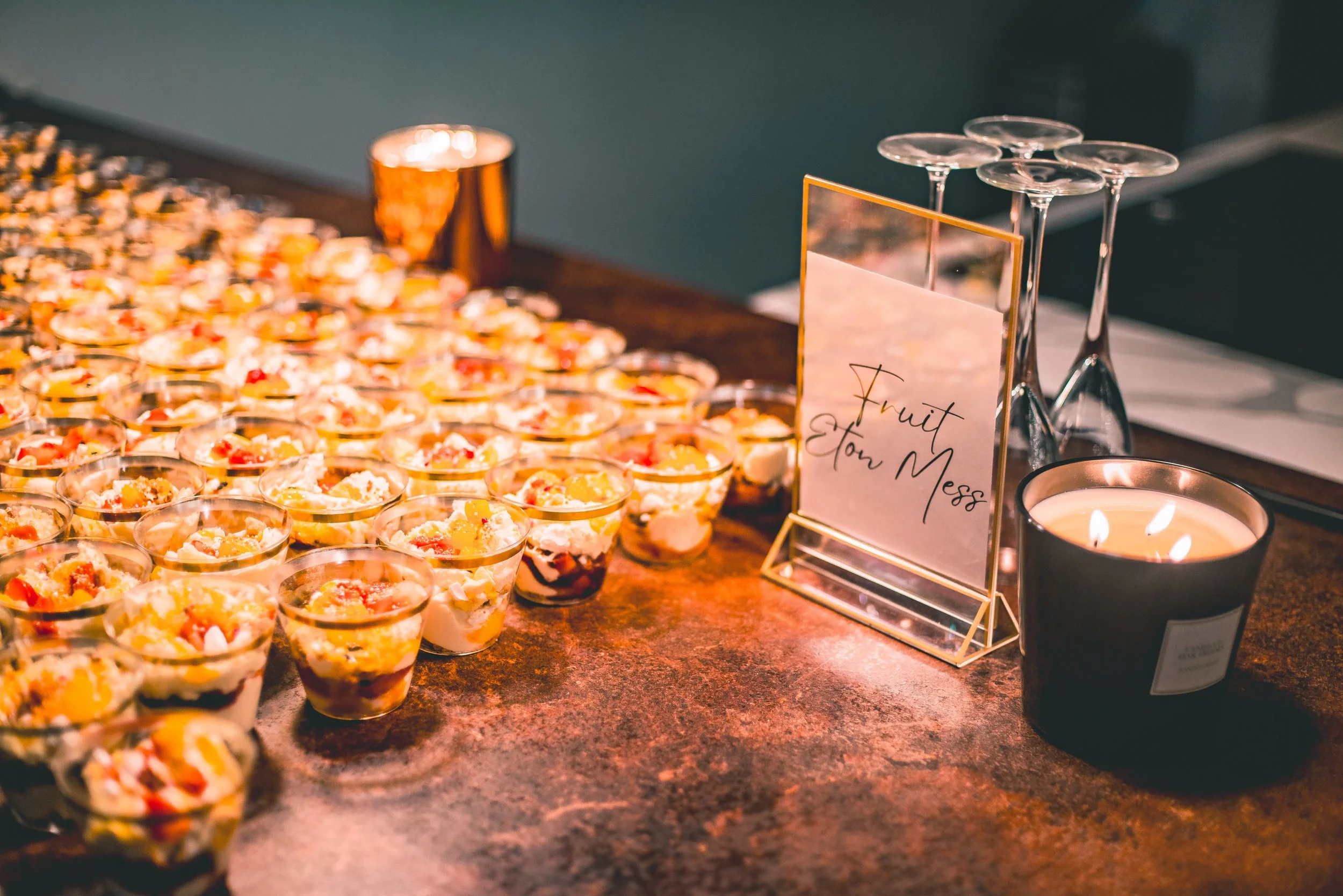 Small dessert cups with fruit toppings arranged on a wooden table, with a candle, a copper-colored cup, and a wine glass holder nearby, and a sign that says 'Fruit on Mess'.