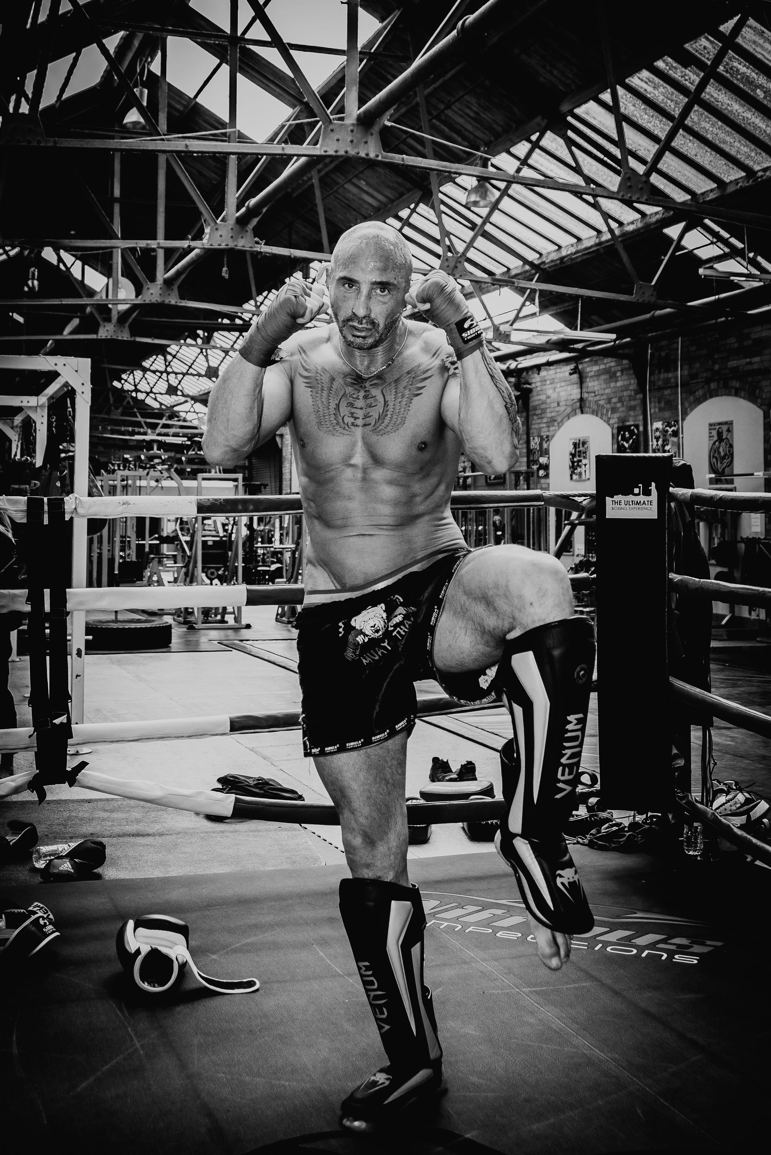 A muscular, shirtless man training in a boxing gym, wearing boxing gloves and shorts, standing inside a boxing ring with gym equipment and graffiti on brick walls in the background.