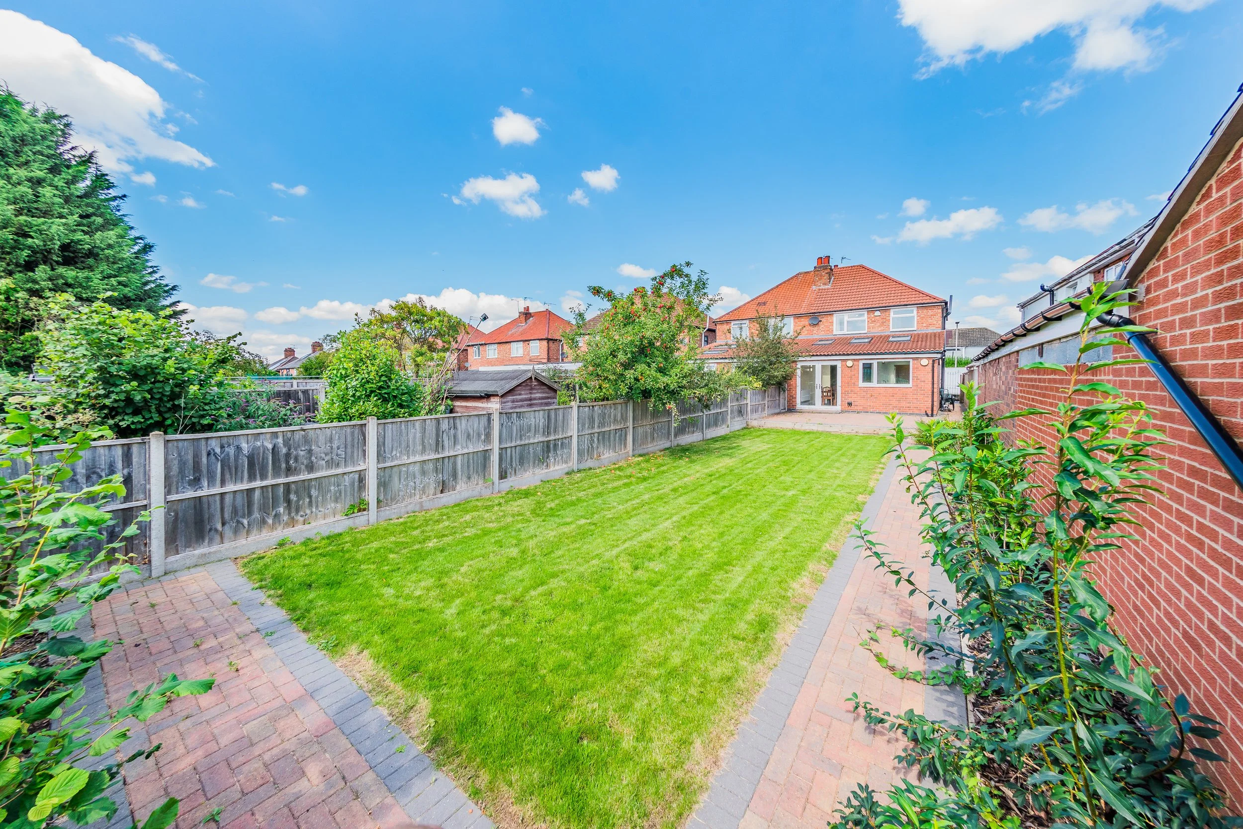 A well-maintained backyard with a green lawn, brick patio, wooden fence, trees, and bushes, with neighboring houses and a blue sky with clouds.