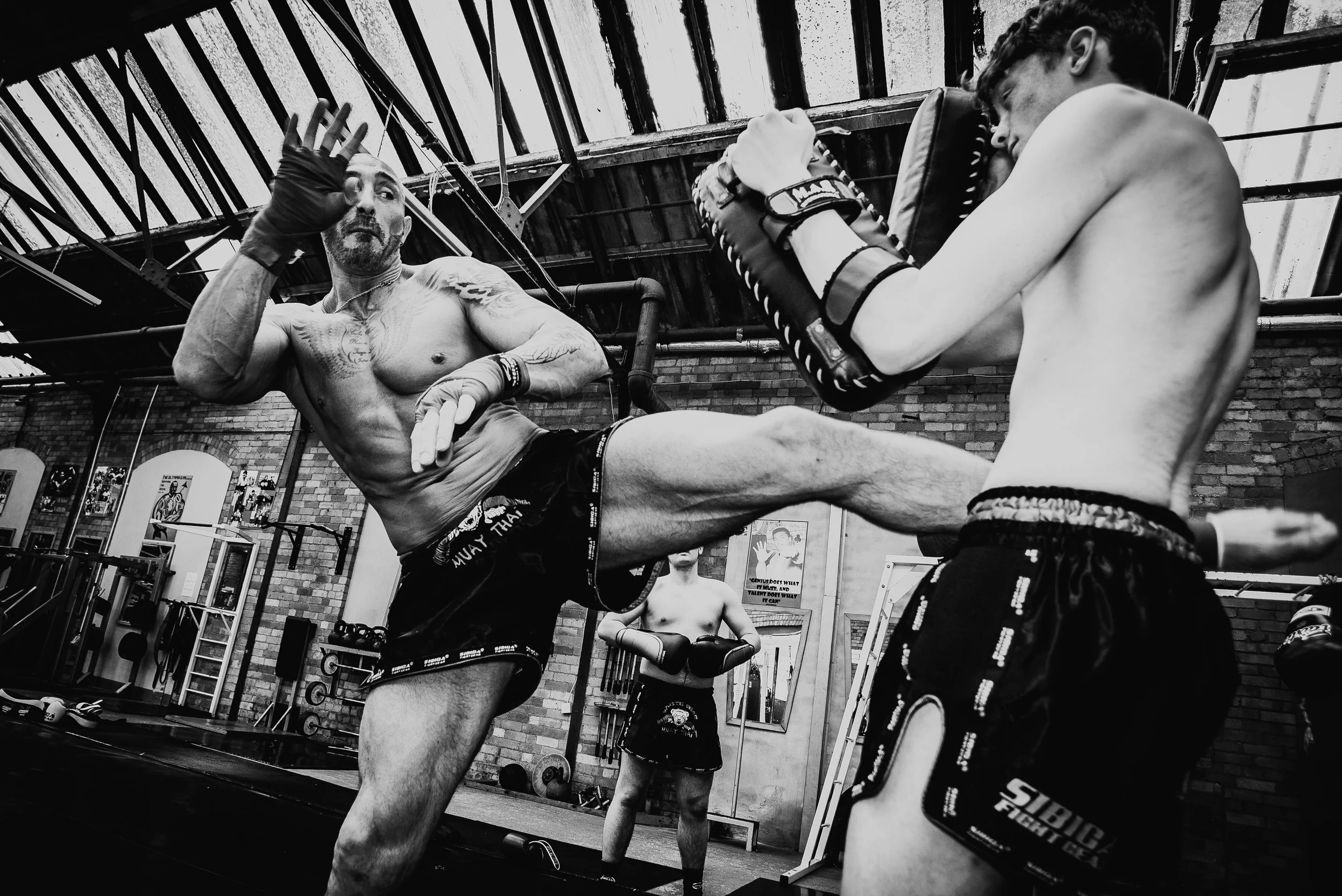 Two shirtless men practicing martial arts in a gym, one delivering a kick to the other, with gym equipment and posters in the background.