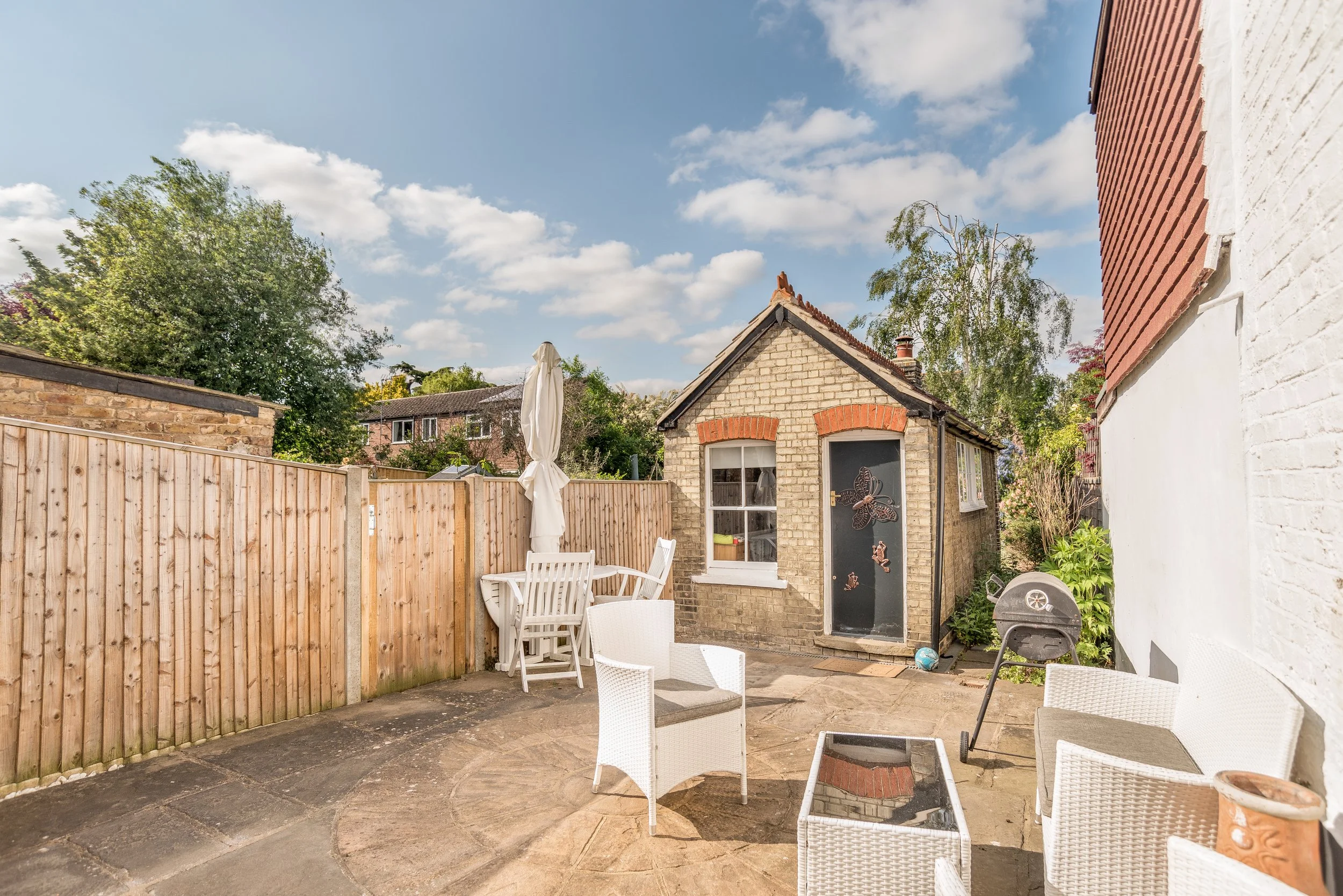 A backyard patio with white outdoor furniture, a small brick building with a butterfly decoration on the door, a wooden fence, and trees under a partly cloudy sky.