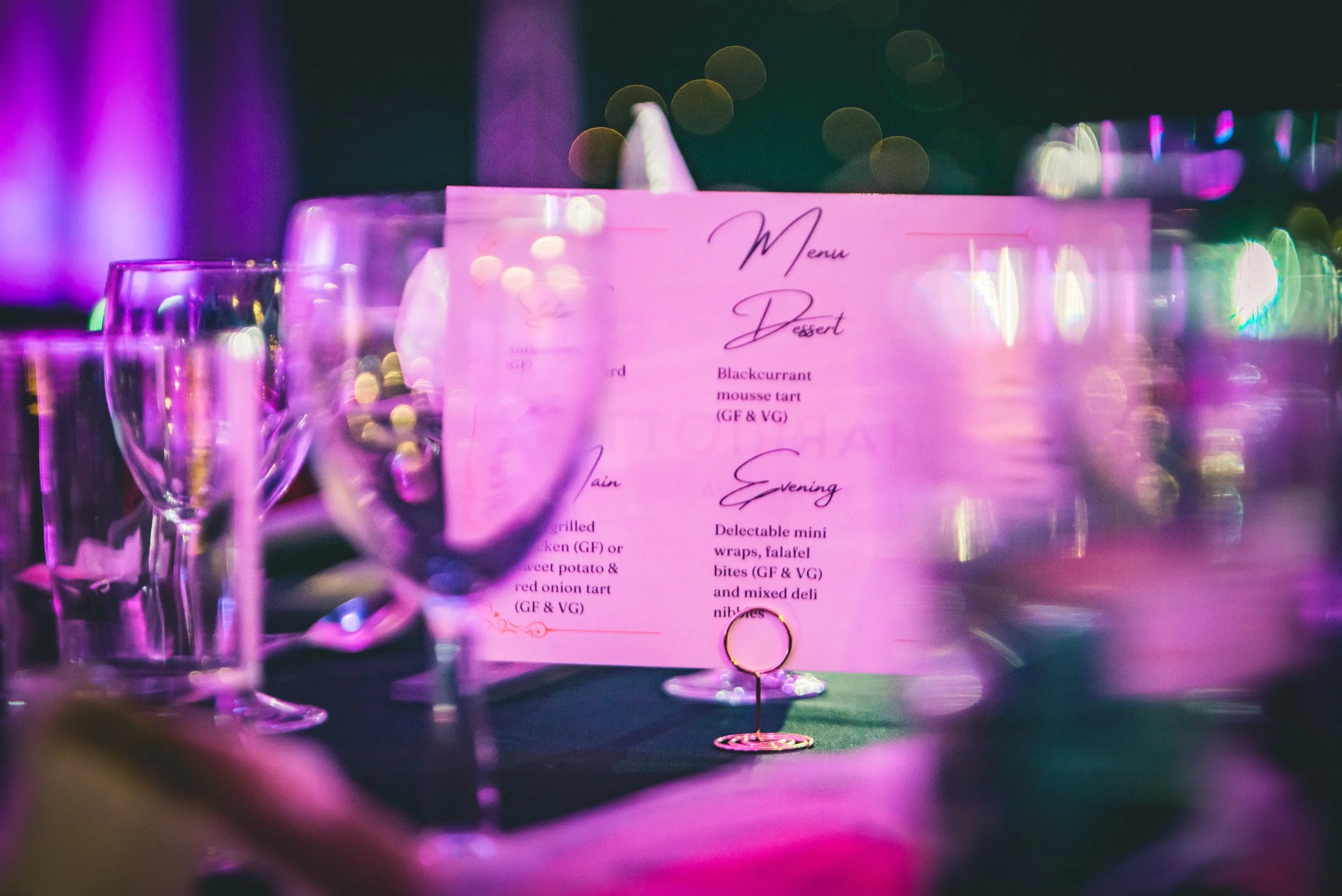A pink menu card on a table with bar glasses and a dim, colorful background at a restaurant or bar.