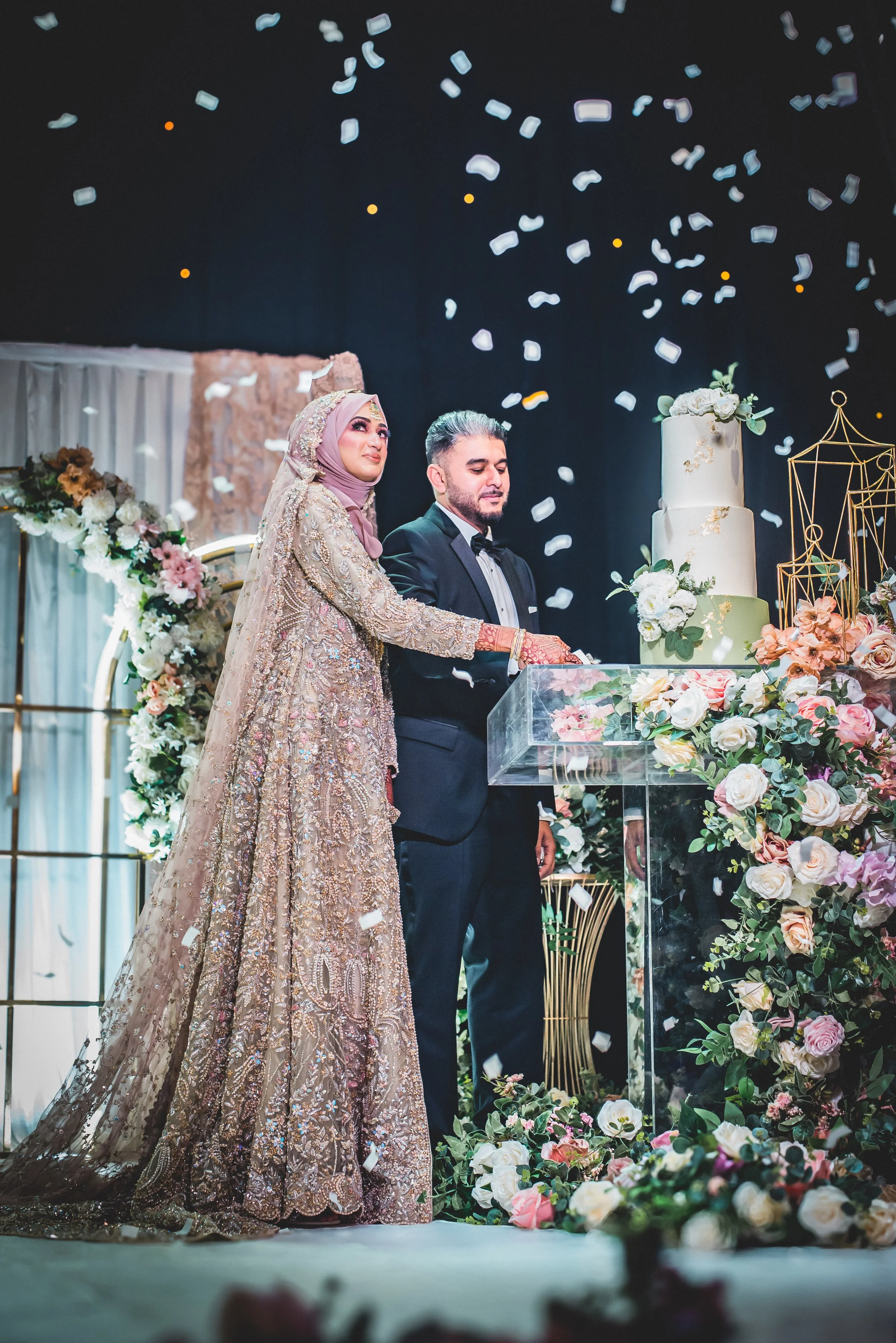 A bride and groom stand together during their wedding reception, holding a cake knife. The bride is dressed in an ornate gold gown with a hijab, and the groom in a tuxedo. White and pink roses decorate the table, and a large, tiered wedding cake is visible. White confetti falls from above, creating a celebratory atmosphere.
