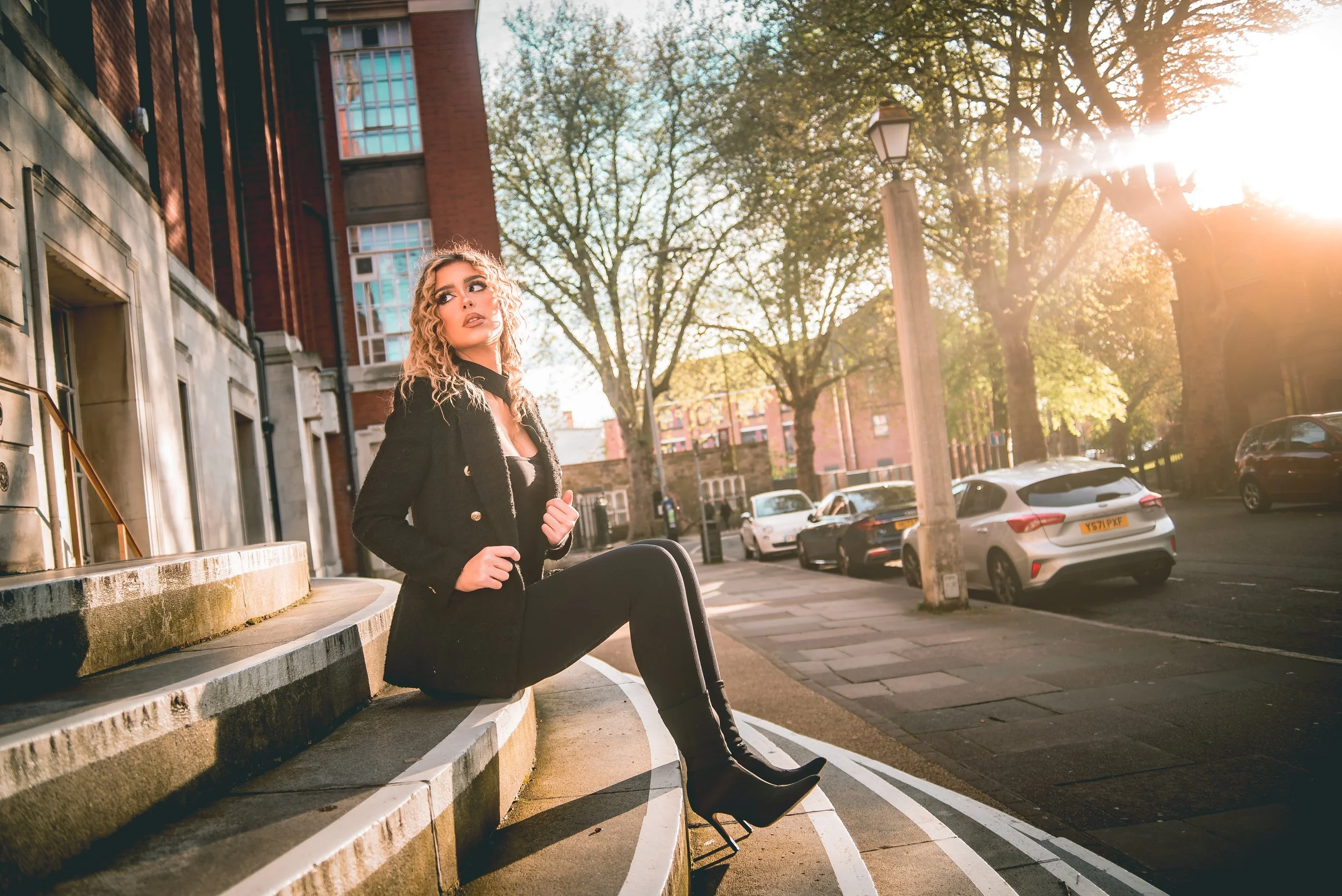 A woman with curly blonde hair sitting on concrete steps in the sunlight, wearing a black coat and boots, with trees and parked cars in the background.