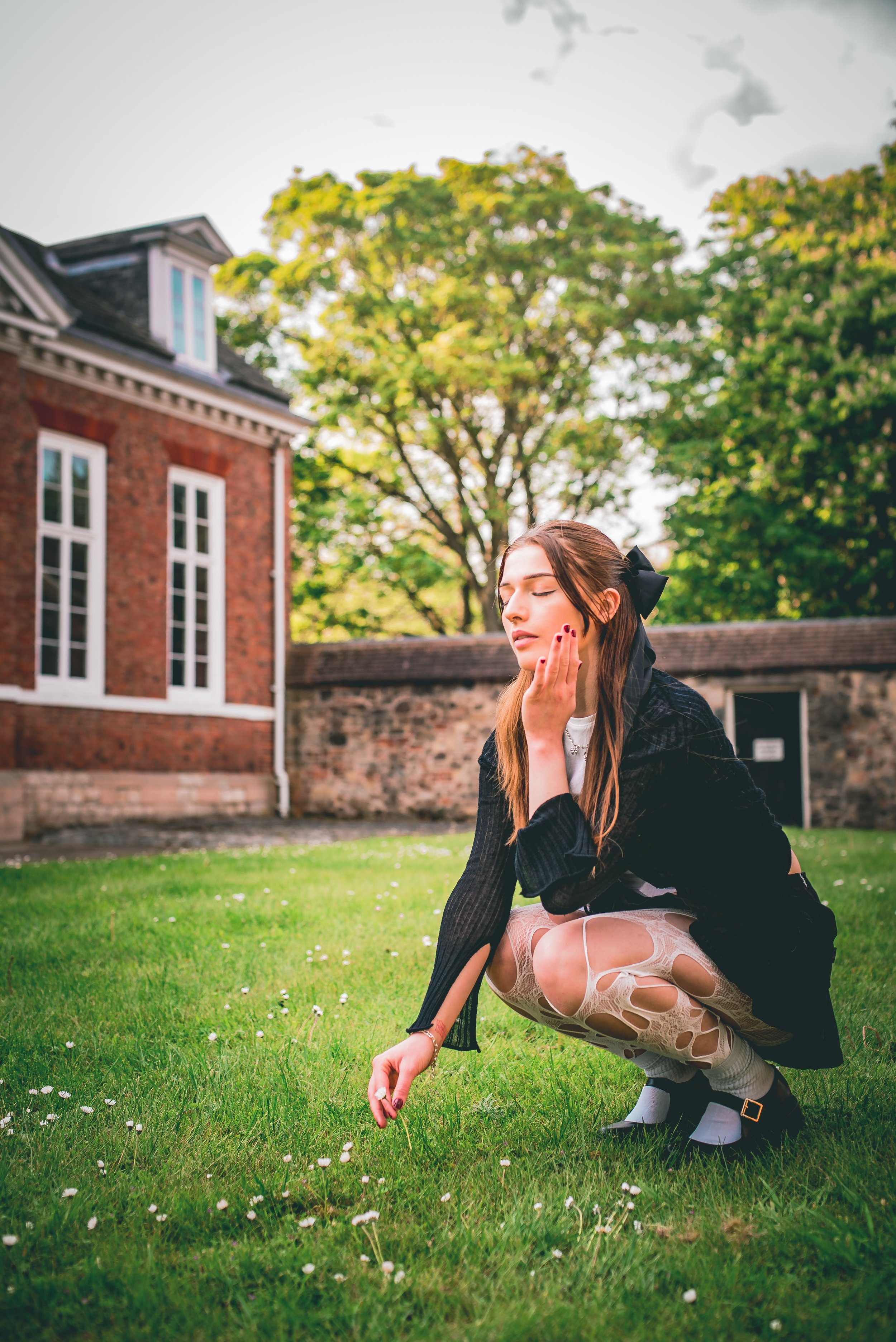 A young woman with long brown hair, wearing a black jacket and ripped jeans, crouches on a green lawn with small white flowers. She is touching her face with one hand and holding a flower with the other. Behind her are trees and a brick building.